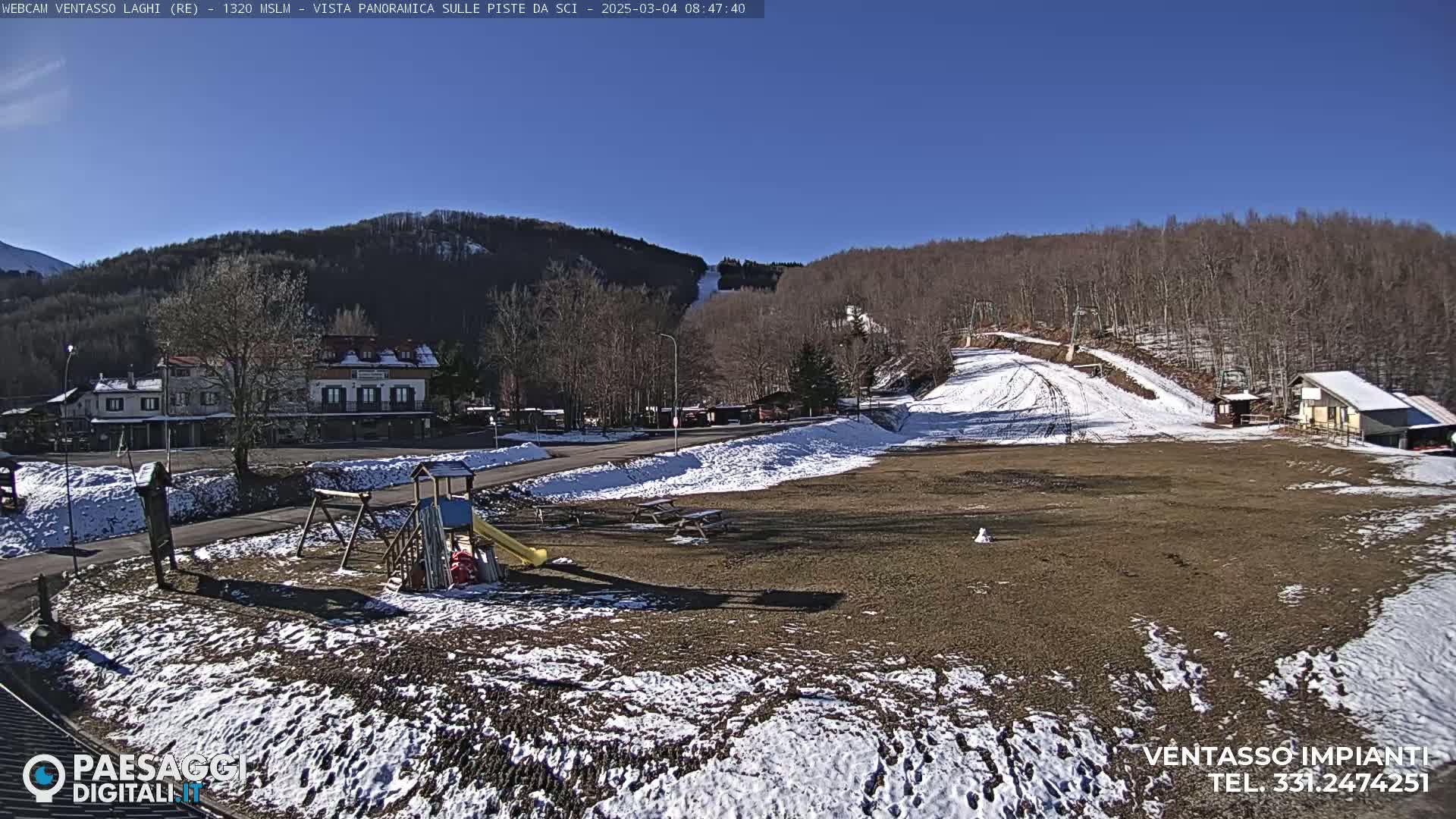 A sunny day reveals a mostly barren ski slope with patches of snow, a playground, a small building, and a larger building further back, all situated in a mountainous area with leafless trees.