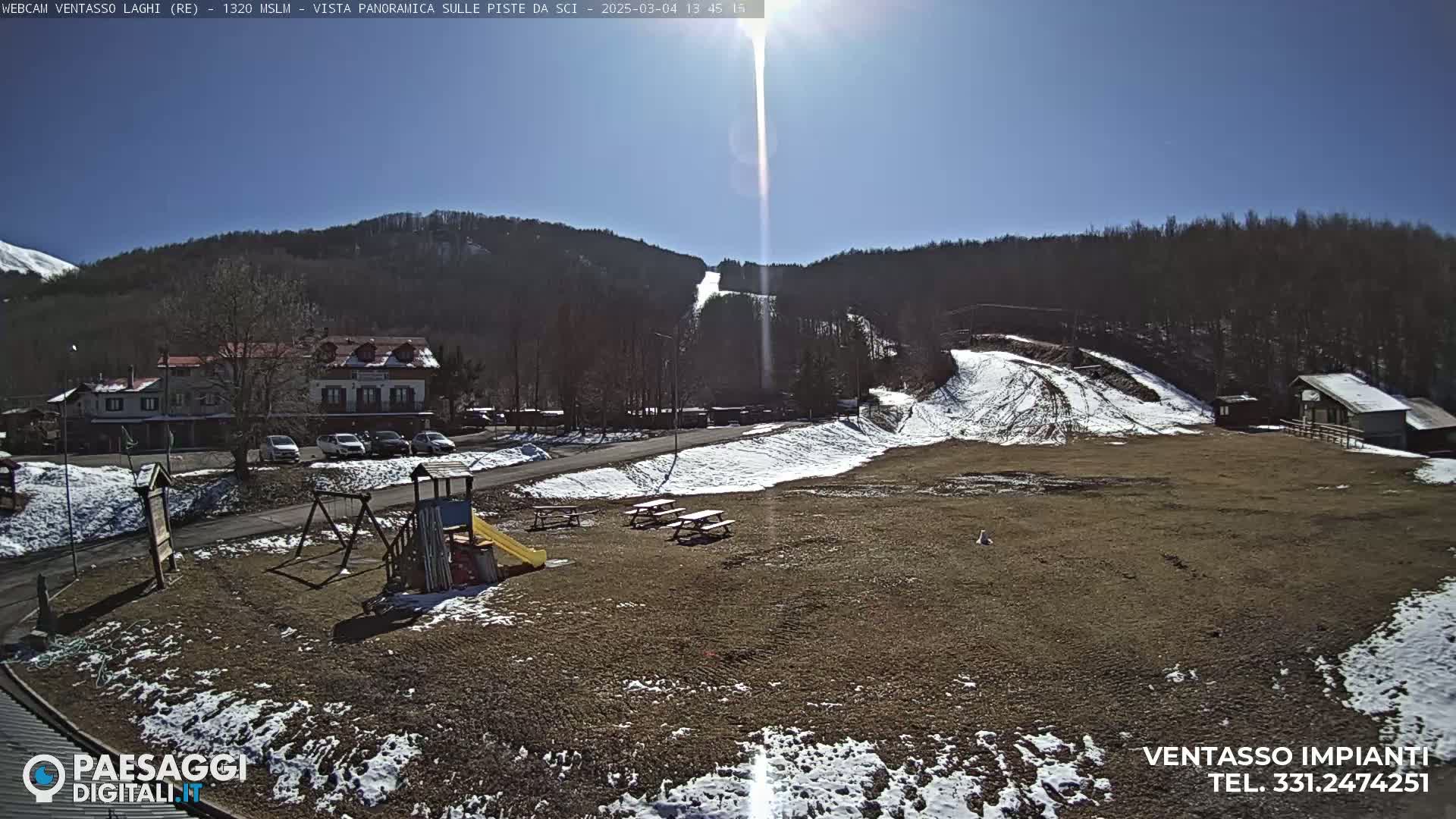 A sunny day at a ski resort shows a playground, picnic tables, a parking lot, buildings, and snowy ski slopes on a hillside in the background.