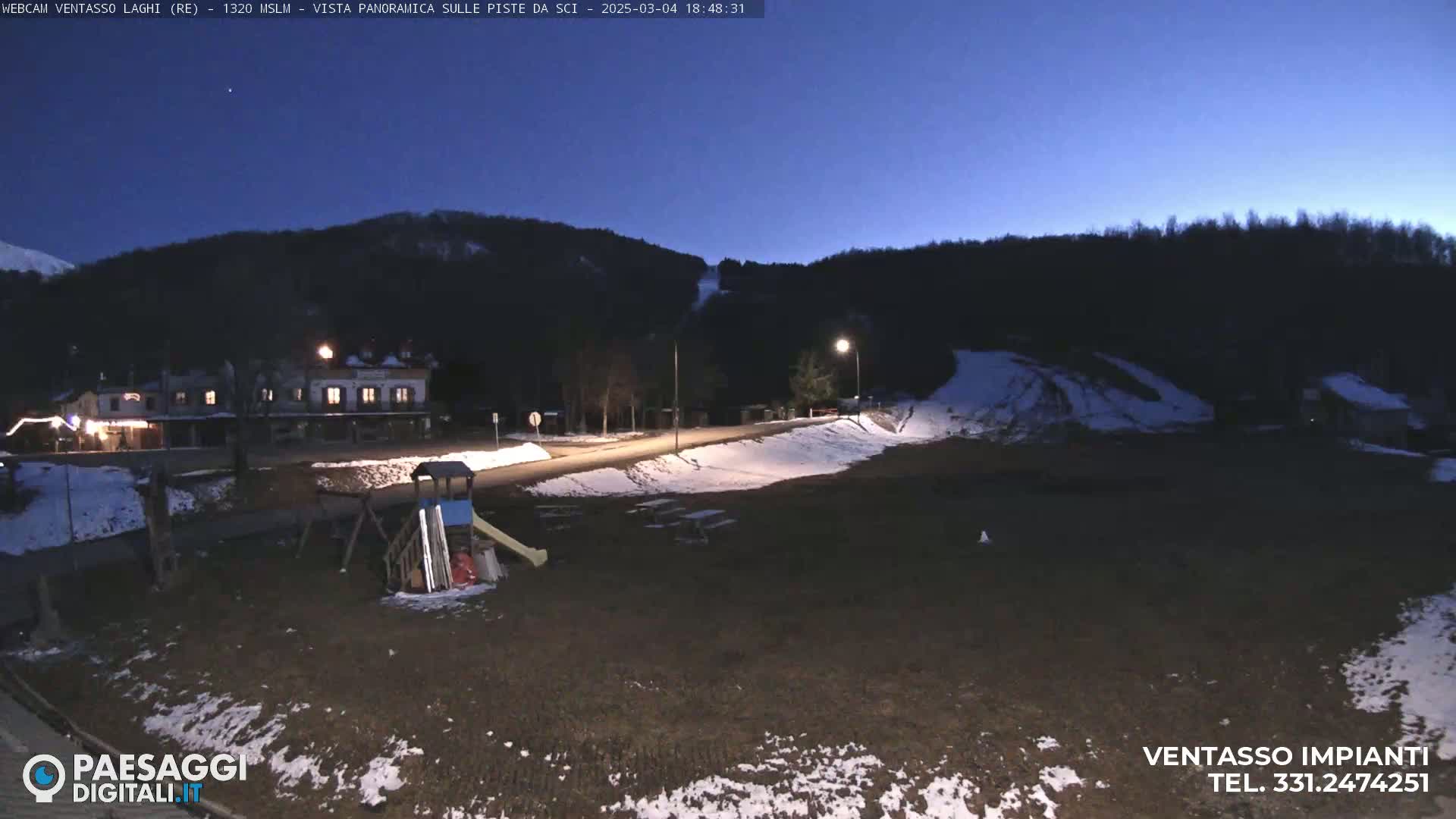 A nighttime view of a mostly empty playground and a small building near a snow-covered hill under a dark blue twilight sky.