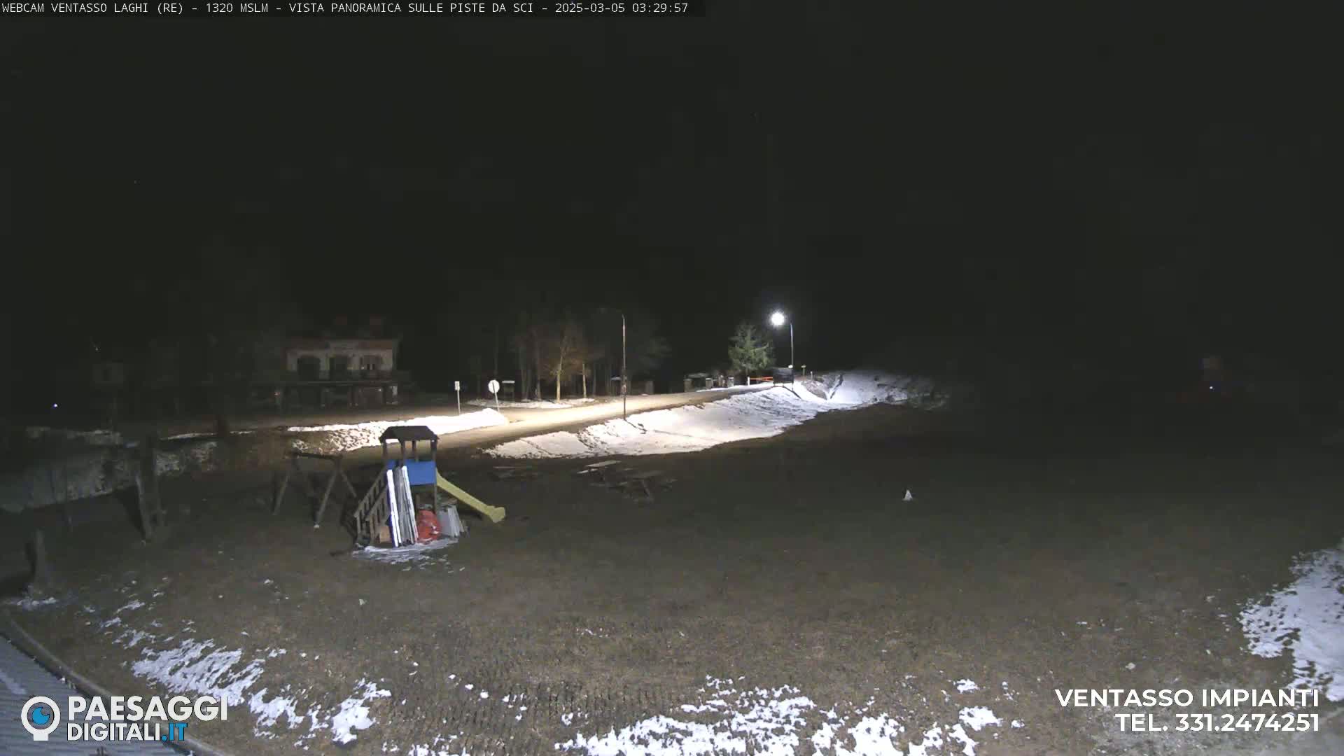 A nighttime view of a mostly empty playground and parking area, lightly dusted with snow, illuminated by artificial lights.