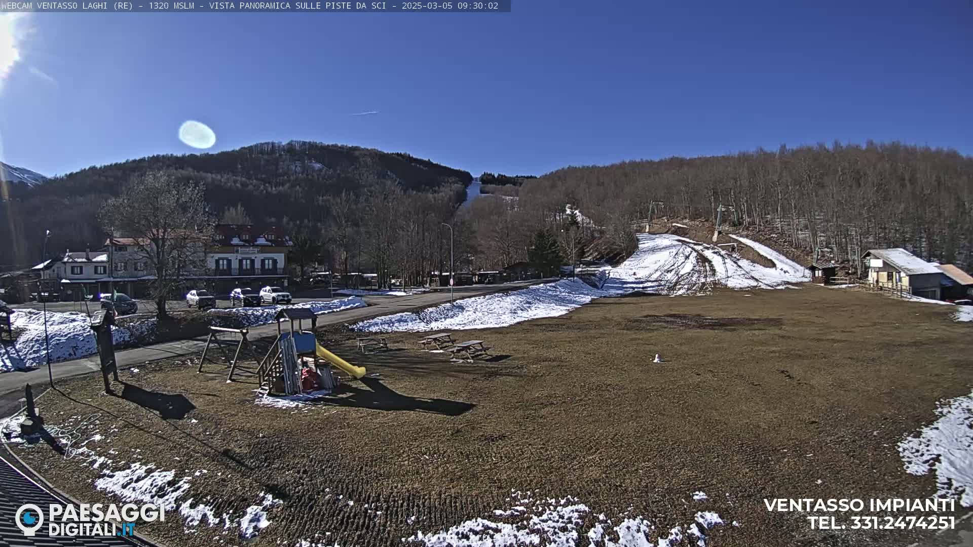 A sunny day reveals a playground, a parking lot, and buildings near a mostly snow-free ski slope with patches of snow in a mountainous area.
