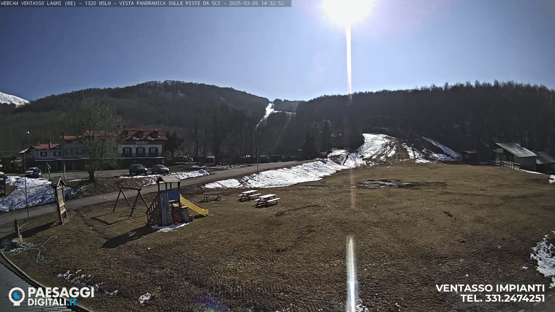 A sunny day reveals a playground, picnic tables, a building, and a ski slope in a mountainous area with patches of snow.