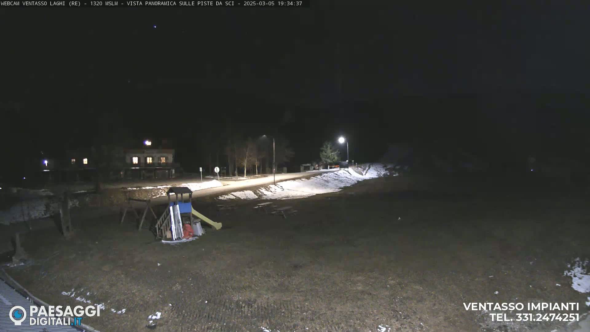 A nighttime view of a playground, a road with some snow, and a building in the distance, under a clear, dark sky.