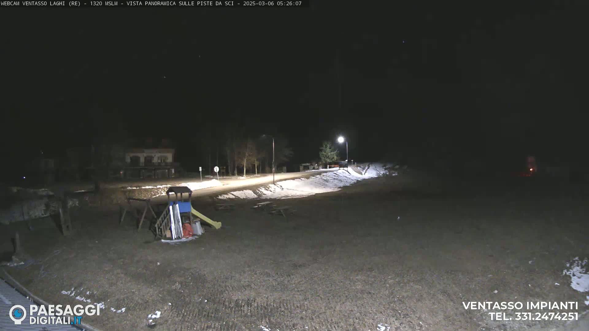A nighttime, snow-dusted playground area with picnic tables and a road is illuminated by streetlights.