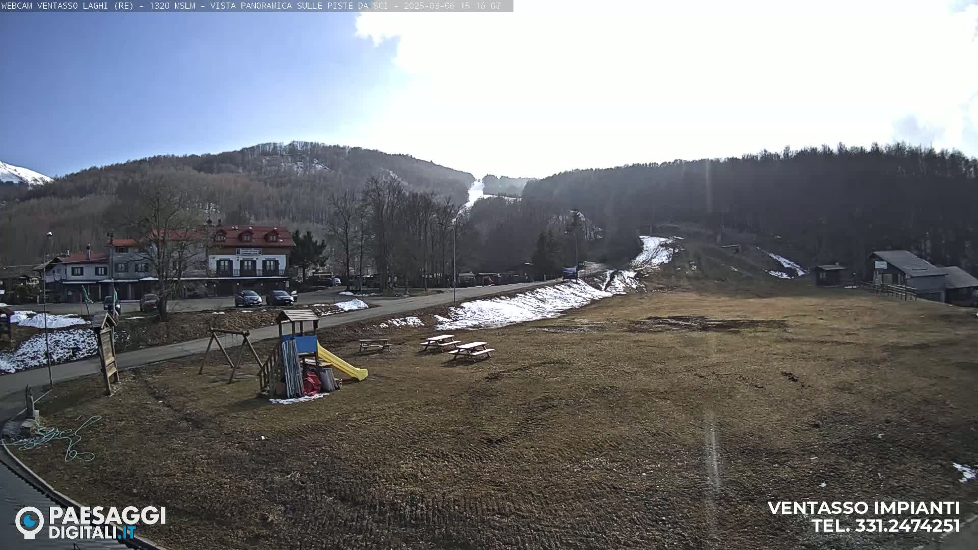 A sunny day reveals a playground, picnic tables, and buildings near a ski slope in a mountainous area with some patchy snow.