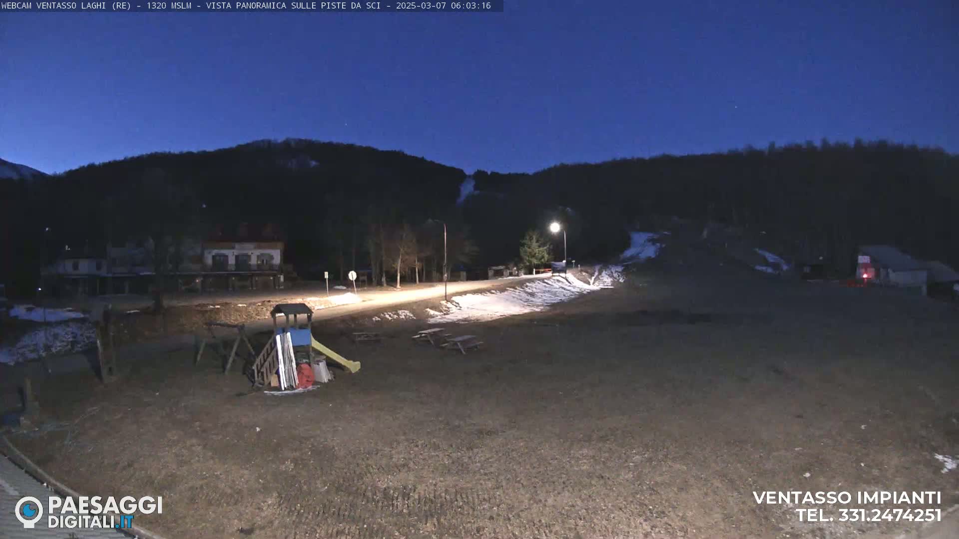 A nighttime view of a mostly empty area with a playground, picnic tables, a road, and buildings in the background under a dark blue, clear sky.