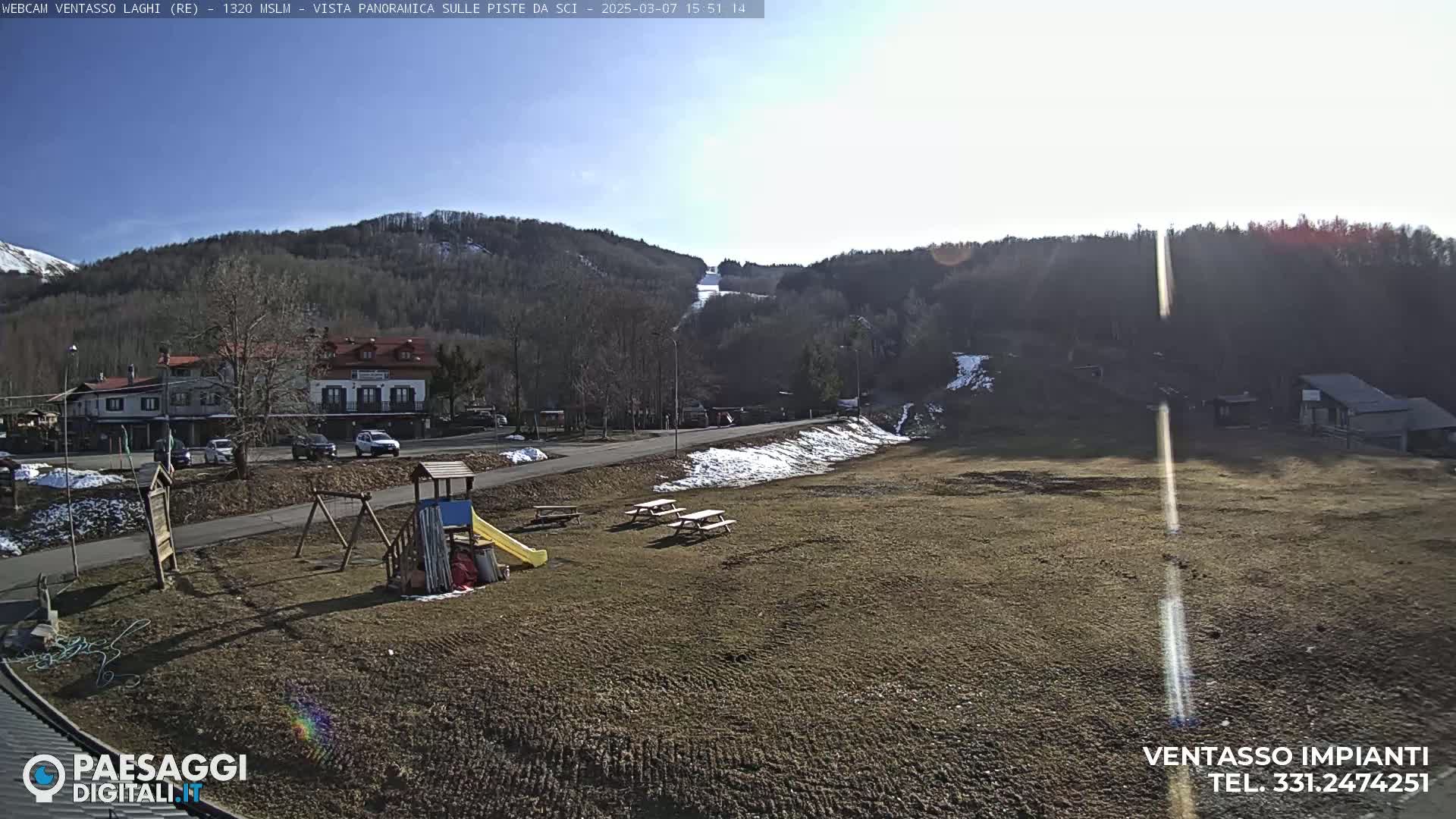 A sunny day reveals a playground, picnic tables, and a road leading to buildings nestled at the base of a snow-dusted mountainside.