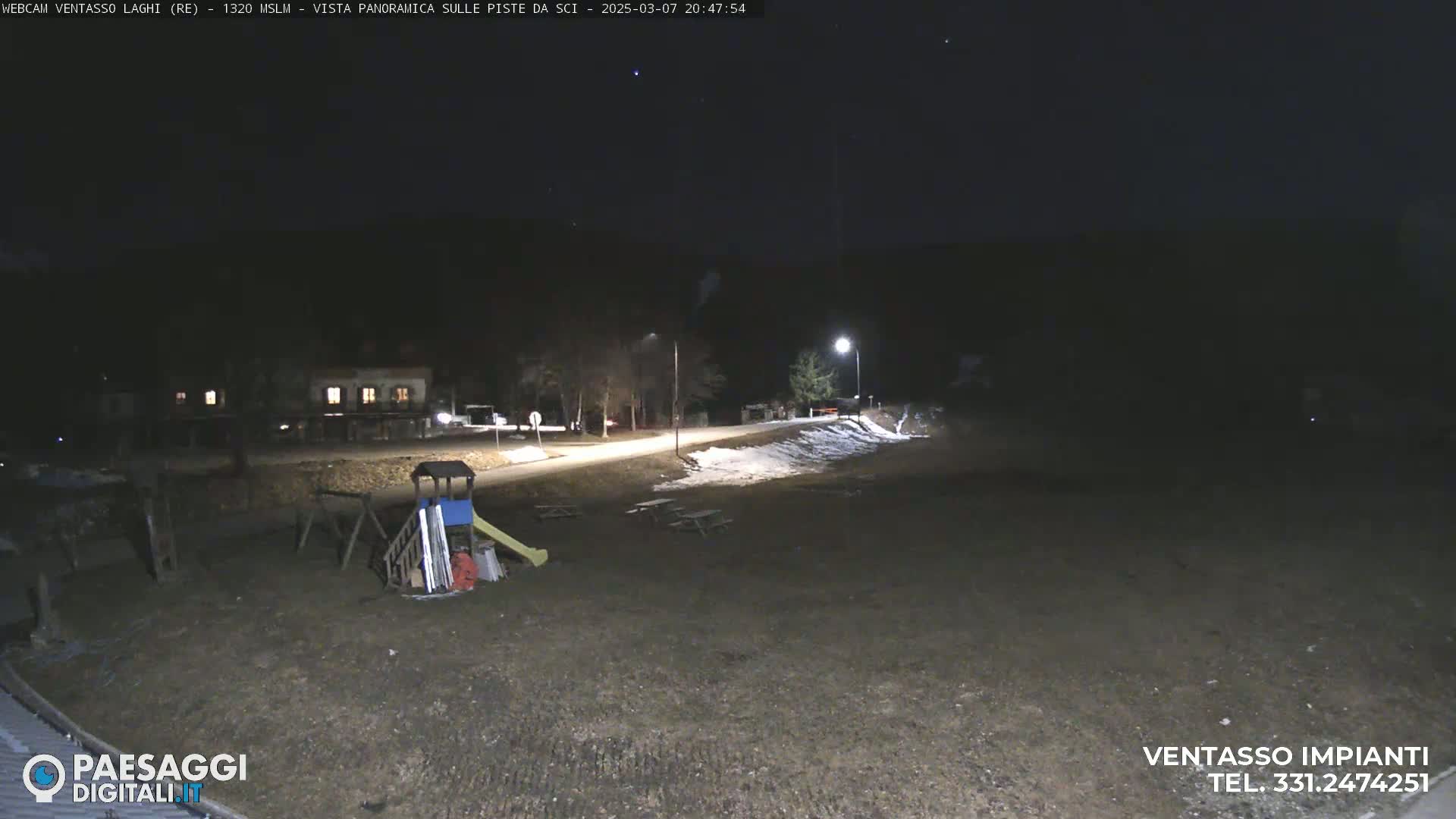 A nighttime view of a mostly empty playground and parking area with some snow on the ground, lit by streetlights under a clear, dark sky.