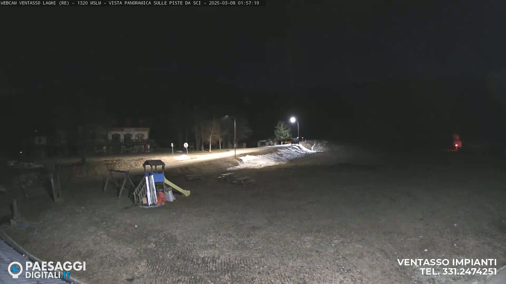 A nighttime, snow-dusted, empty playground area is illuminated by a streetlight, with a road and a few buildings visible in the distance.