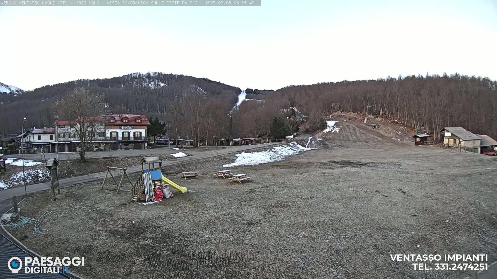 A playground and picnic tables are situated in a gravel area overlooking a ski slope and a village nestled at the foot of a sparsely wooded hill, under a mostly clear sky.