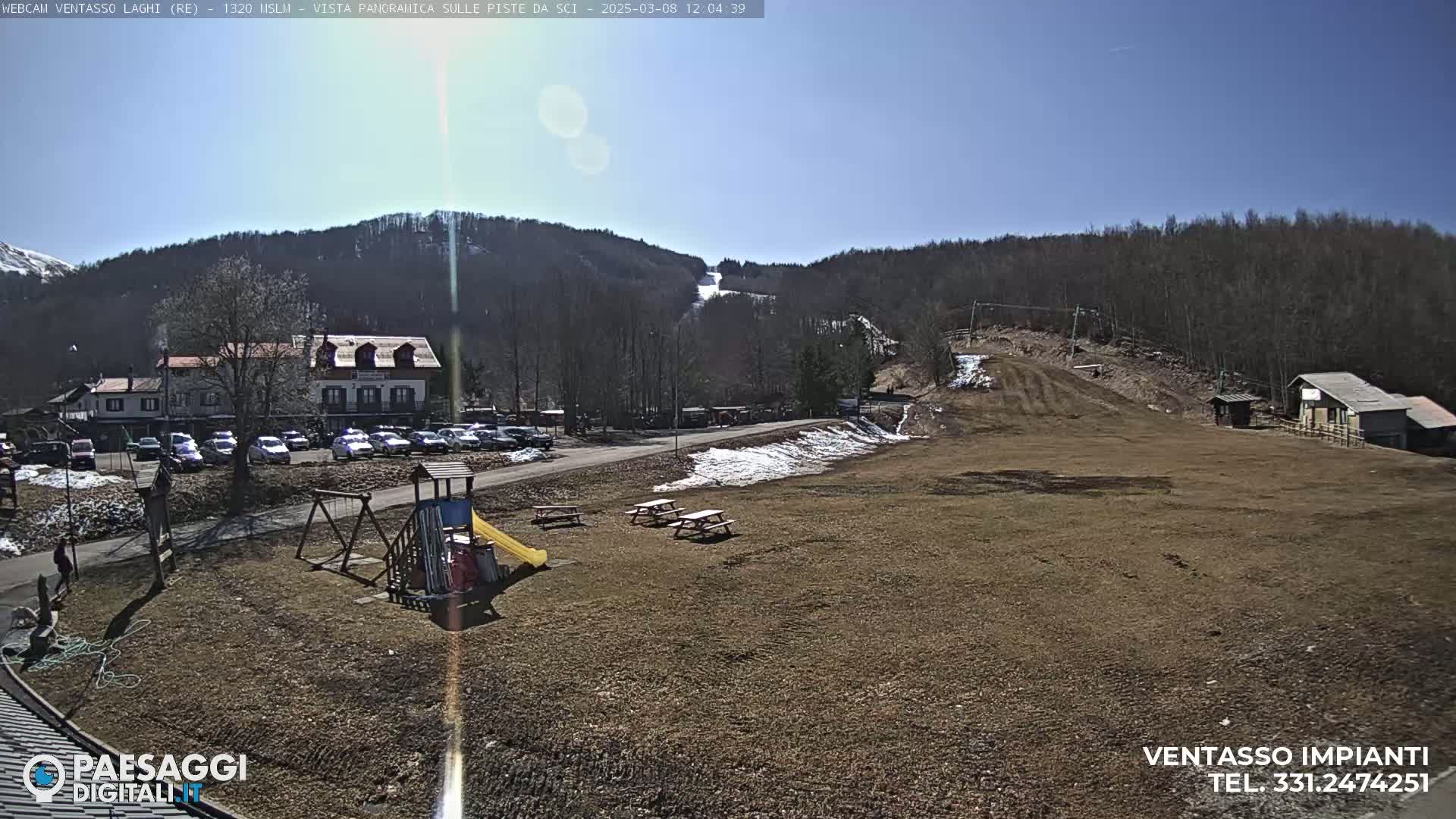 A sunny day reveals a ski slope with a few patches of snow, a parking lot full of cars, a playground, picnic tables, and buildings nestled in the foothills of a mountain range.
