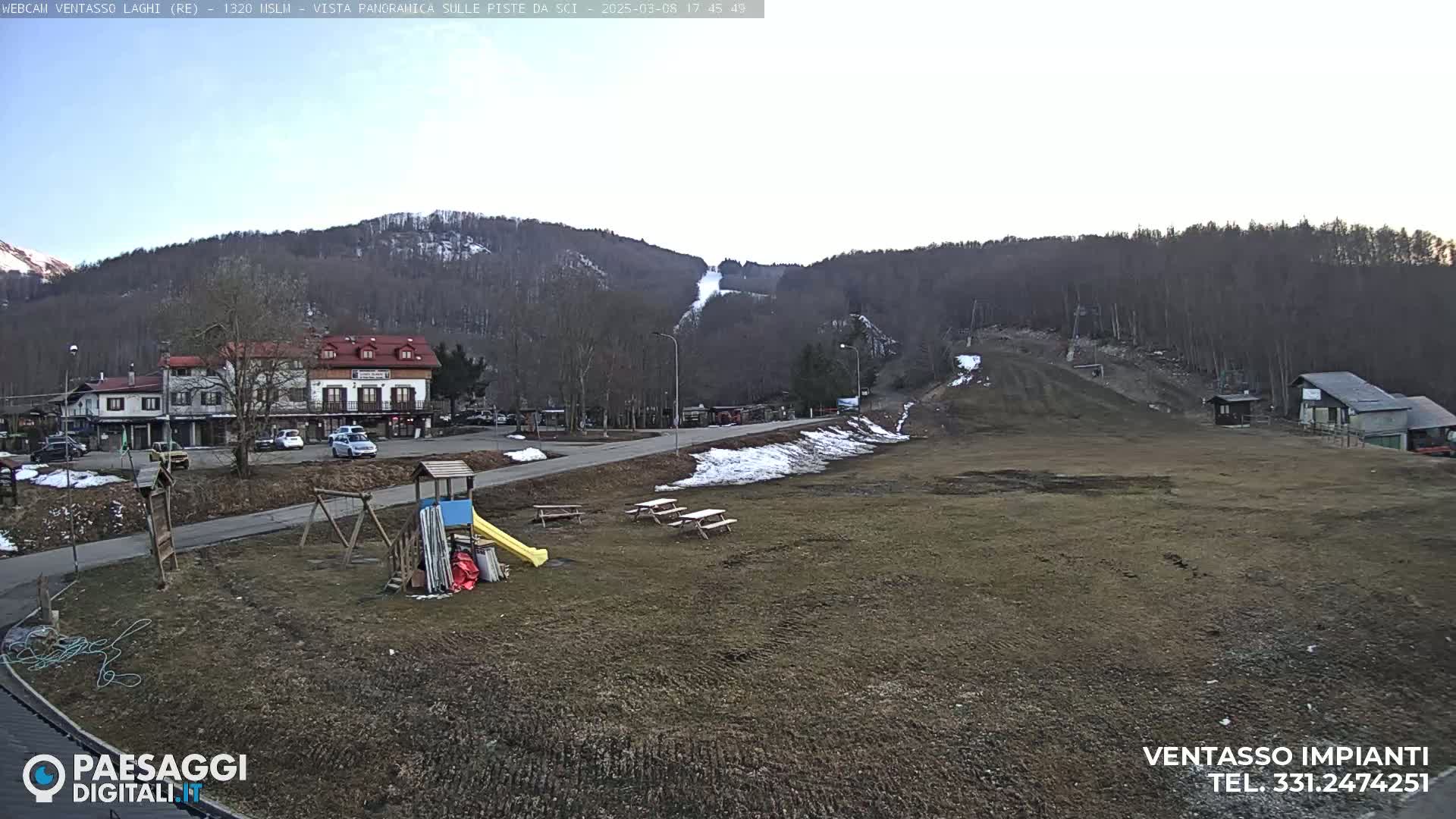 A mostly clear day reveals a ski slope, a playground, a few buildings, and a parking lot nestled amongst bare, brown hills.