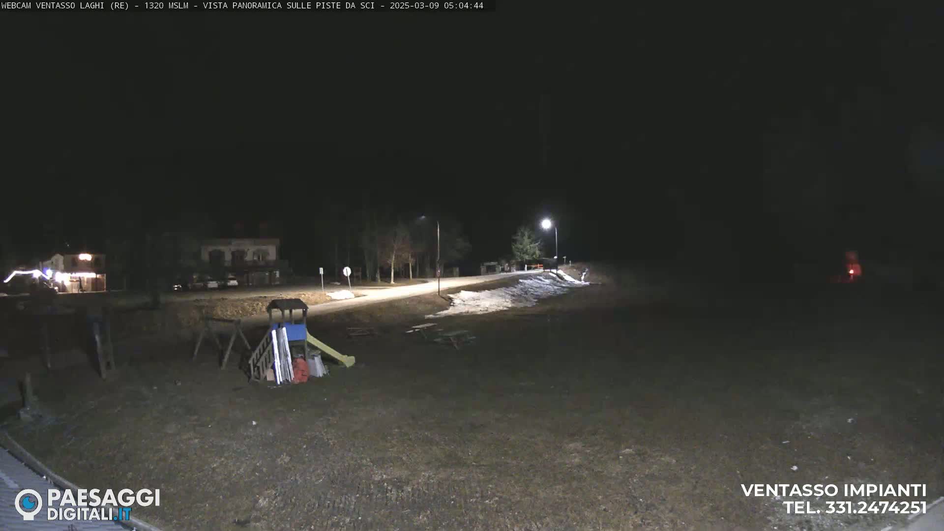 A nighttime view of a mostly empty playground and a road with patches of snow, illuminated by streetlights.