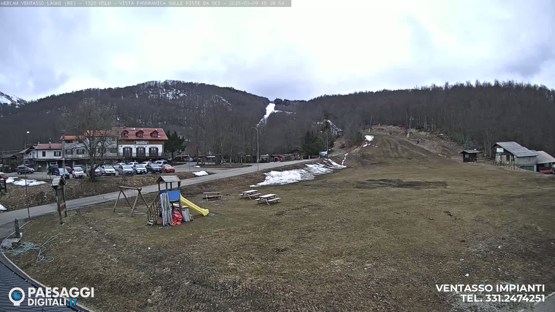 A playground and picnic tables sit in a grassy area below a ski slope on a mostly cloudy day, with a building and parked cars visible in the background.