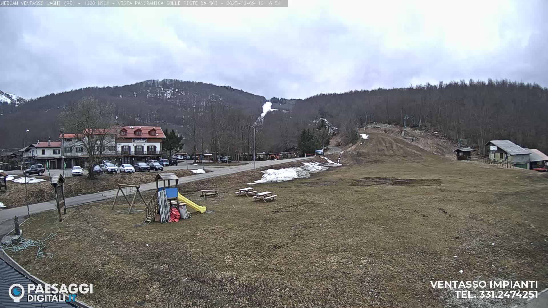 A mostly brown, grassy area with a playground, picnic tables, a parking lot, and buildings sits at the base of a mostly barren, snow-dusted mountain under a cloudy sky.