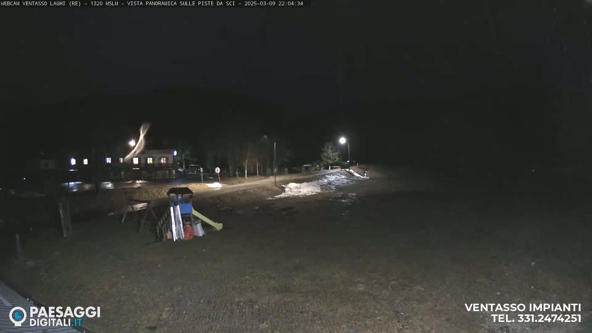 A nighttime view of a mostly empty paved area with some snow patches, a building in the distance, and playground equipment nearby.