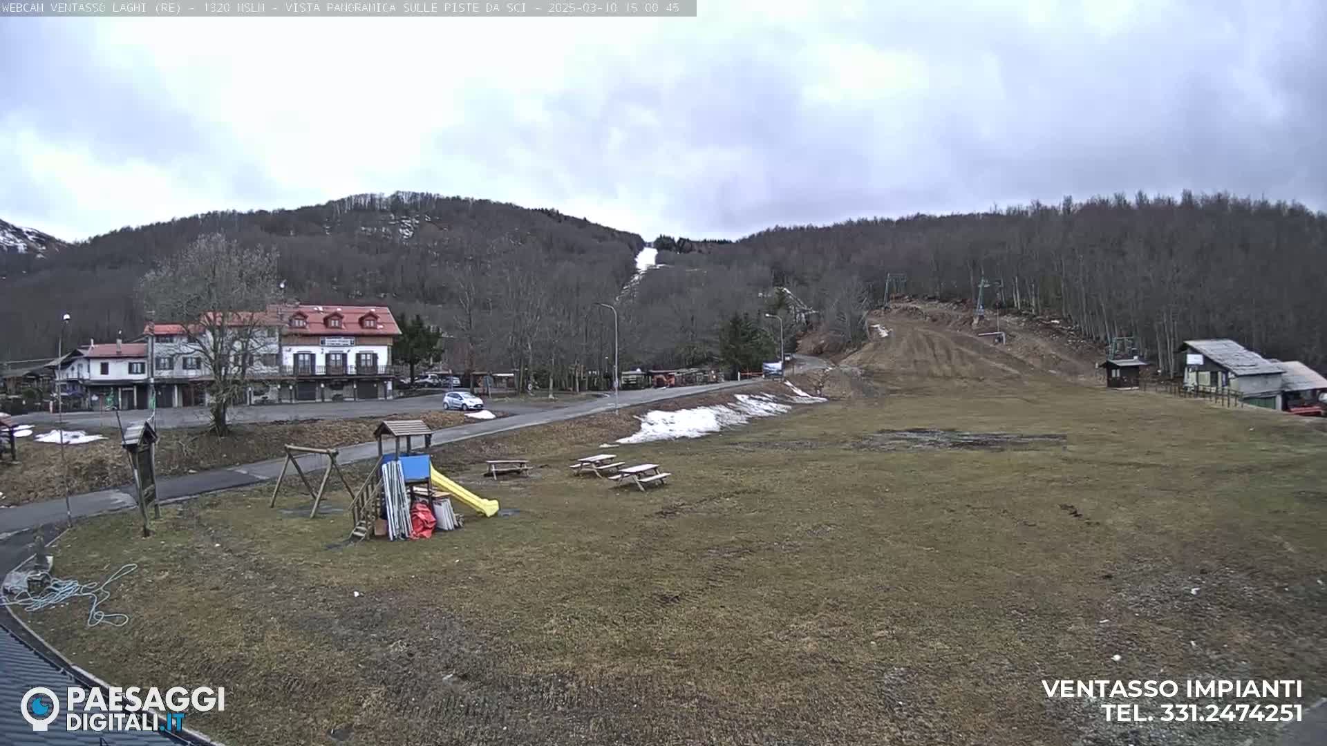 A mostly clear, overcast day reveals a view of a ski slope with some snow, a small village with buildings, a playground, and picnic tables in the foreground.