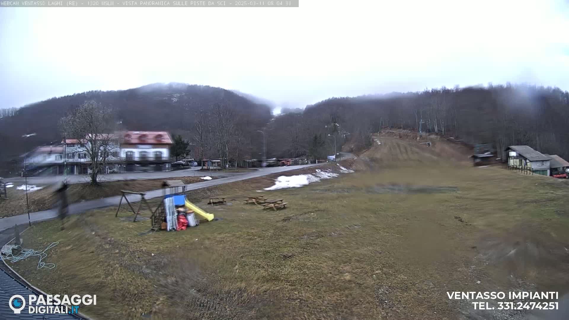 A mostly cloudy day reveals a view of a ski resort with a building, playground, picnic tables, and a ski slope in the background.