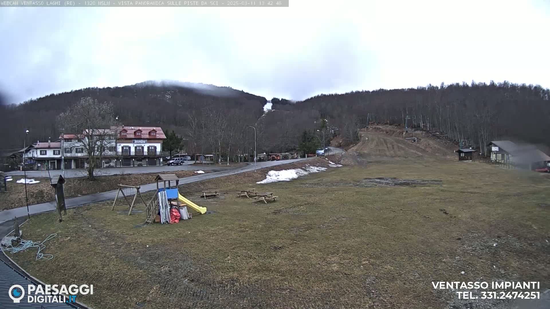 A mostly cloudy day reveals a ski slope with some snow, a playground, and several buildings situated near a wooded hillside.