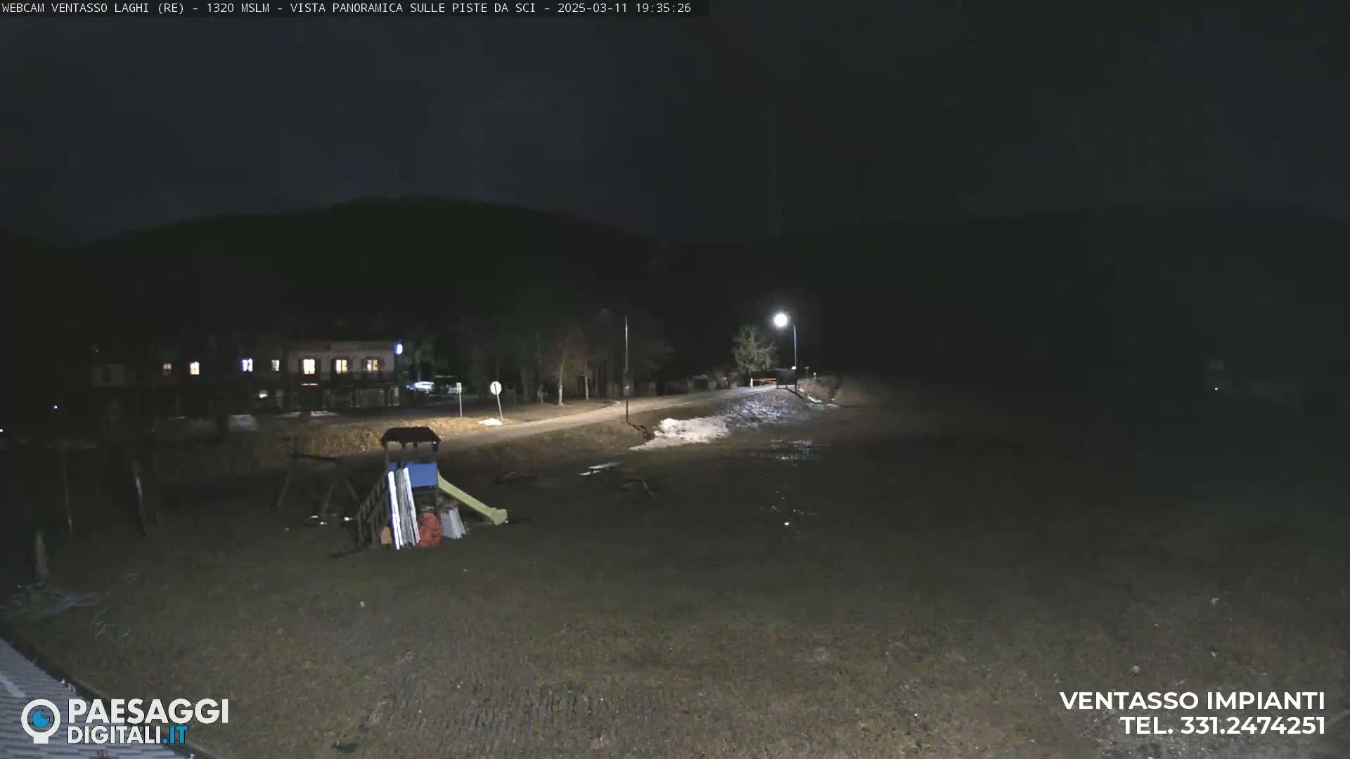 A nighttime view of a mostly empty playground and a road, with a building and some trees visible in the background under a dark sky.