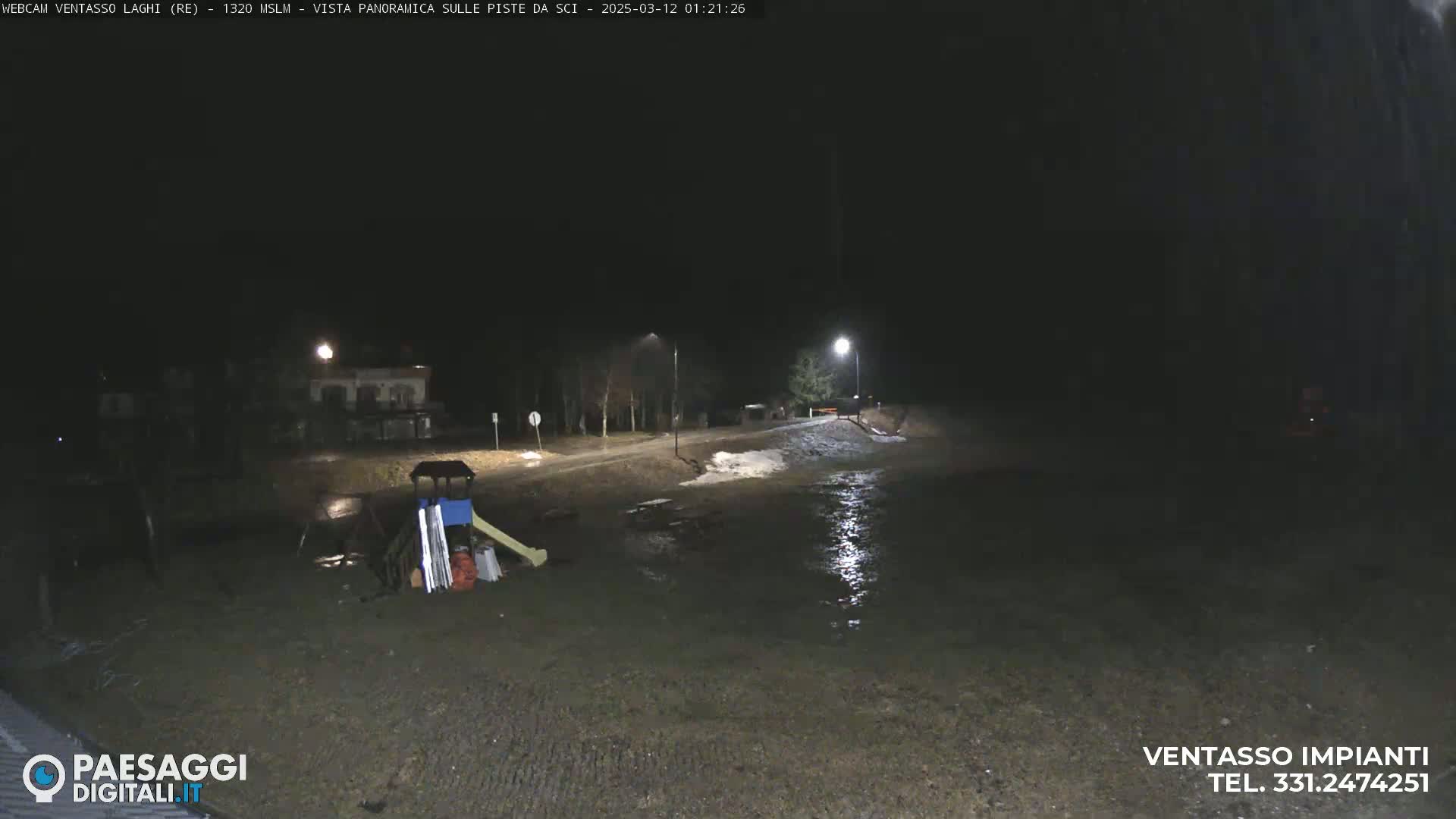 A nighttime view of a flooded playground and road, illuminated by streetlights, with some snow visible in the background.