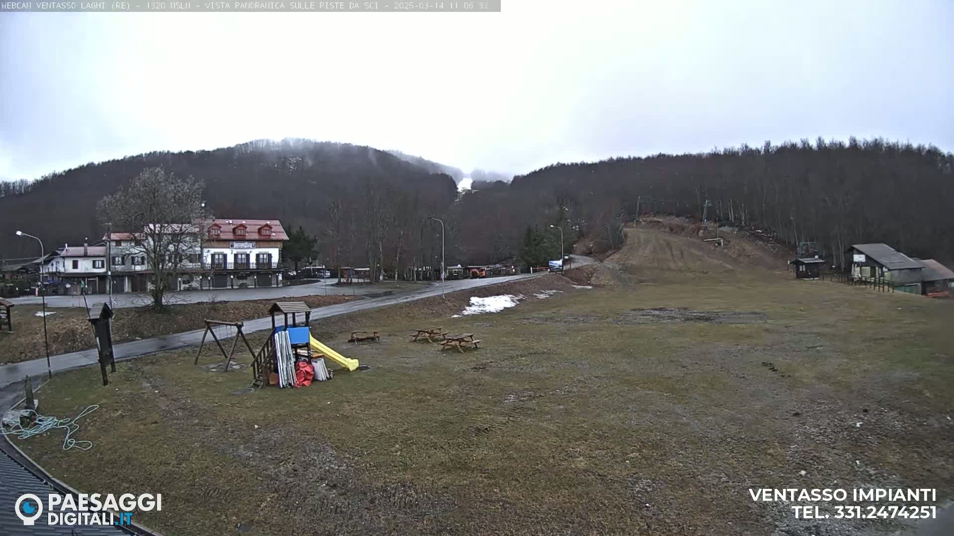 A mostly overcast view of a ski resort area with a playground, picnic tables, and buildings, situated at the base of a hill covered in bare trees.
