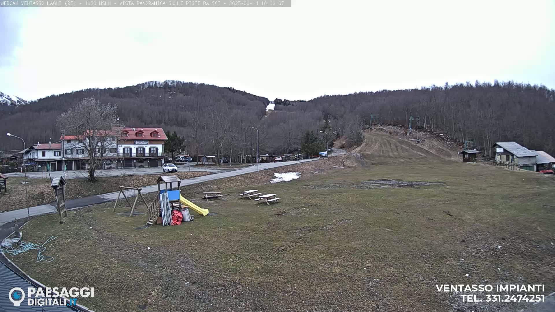 A playground and picnic tables sit in a grassy area near a road and buildings, with a ski slope and bare trees visible on a hill in the background under an overcast sky.