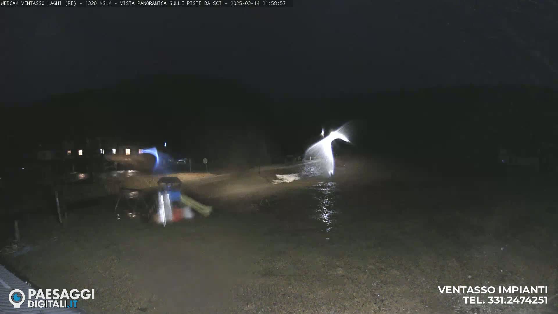At night, under dark skies, a brightly lit area shows a body of water with what appears to be snowmaking equipment operating near a road.