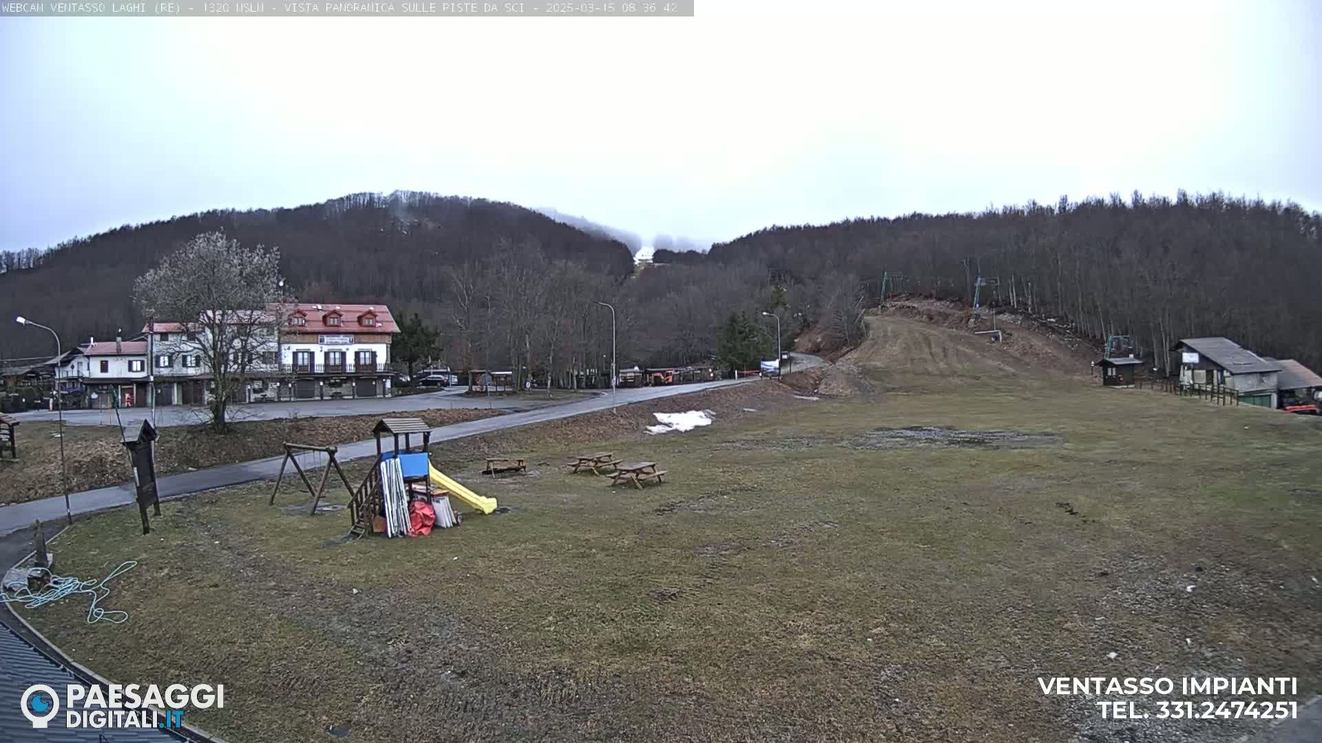 A mostly cloudy day shows a view of a ski slope with a few buildings, a playground, and picnic tables in the foreground.