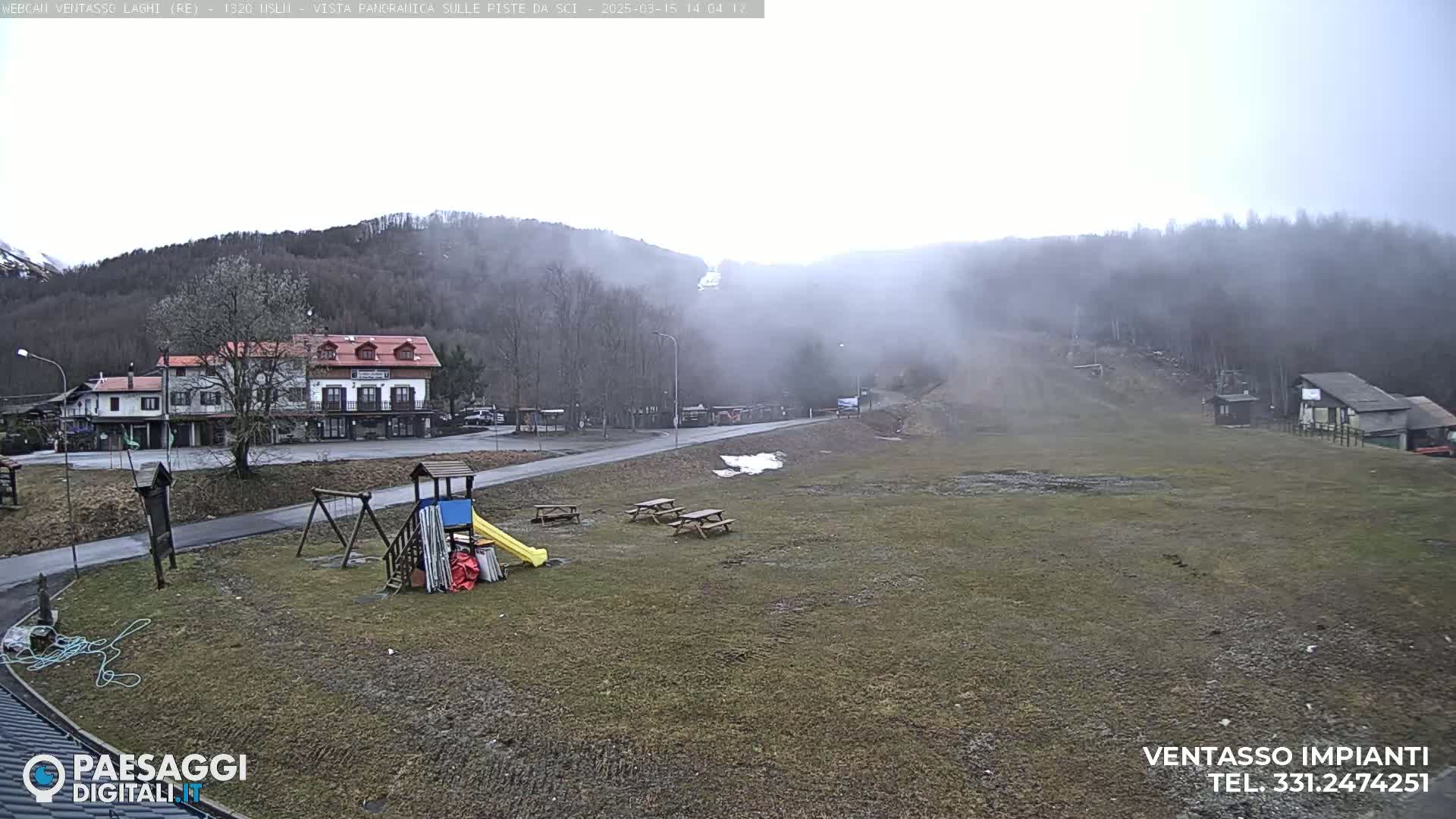 A foggy, overcast day reveals a playground, picnic tables, a building, and a sparsely vegetated area leading up to a misty hillside.
