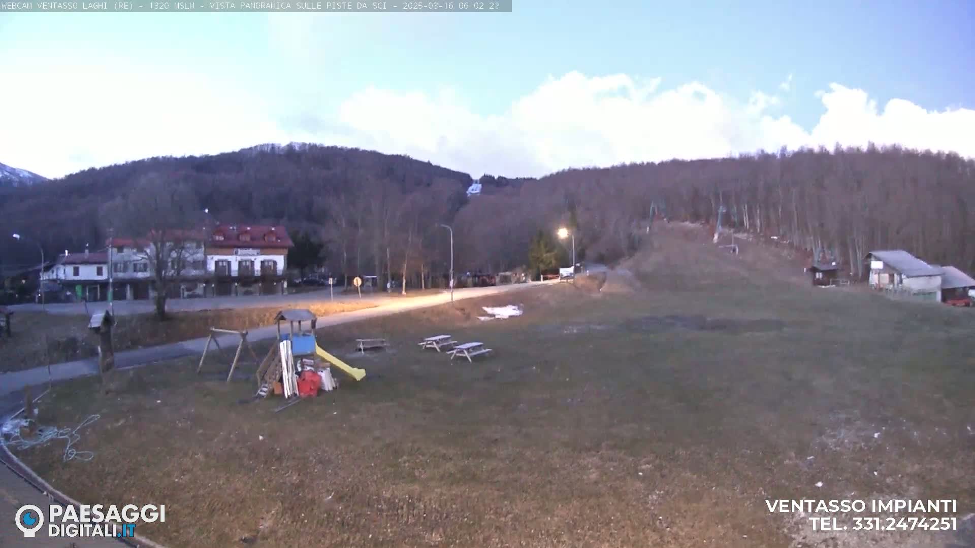 A mostly cloudy, twilight scene shows a playground, picnic tables, buildings, and a sparsely vegetated hillside with a ski lift in the distance.