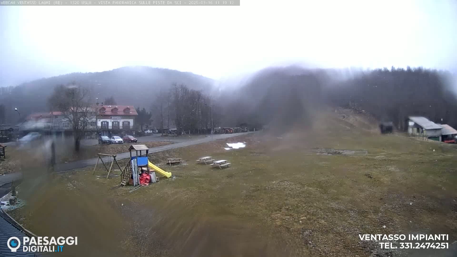 A playground with a slide and swings sits near a building and parking lot in a lightly snowing, foggy landscape with hills in the background.