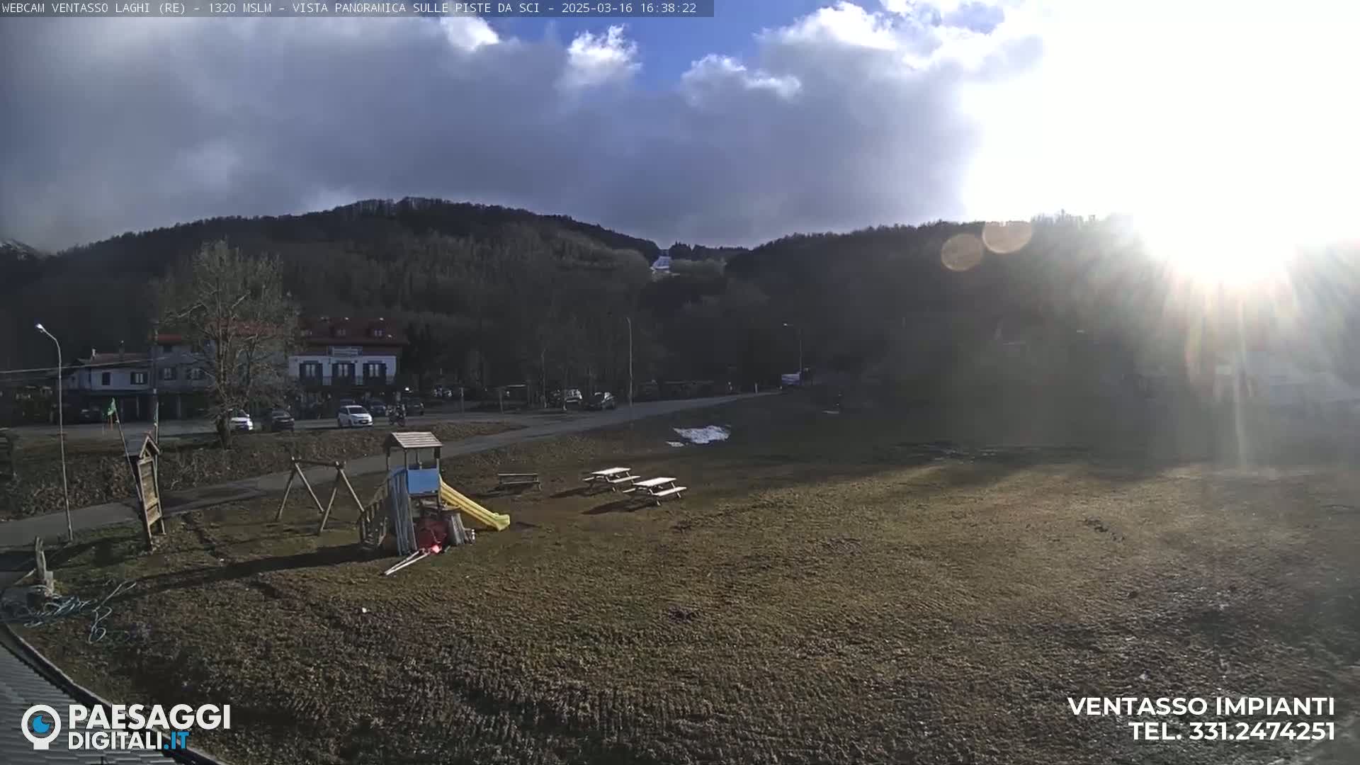 A sunny day reveals a playground, picnic tables, and a few parked cars near a hillside village, with partly cloudy skies.