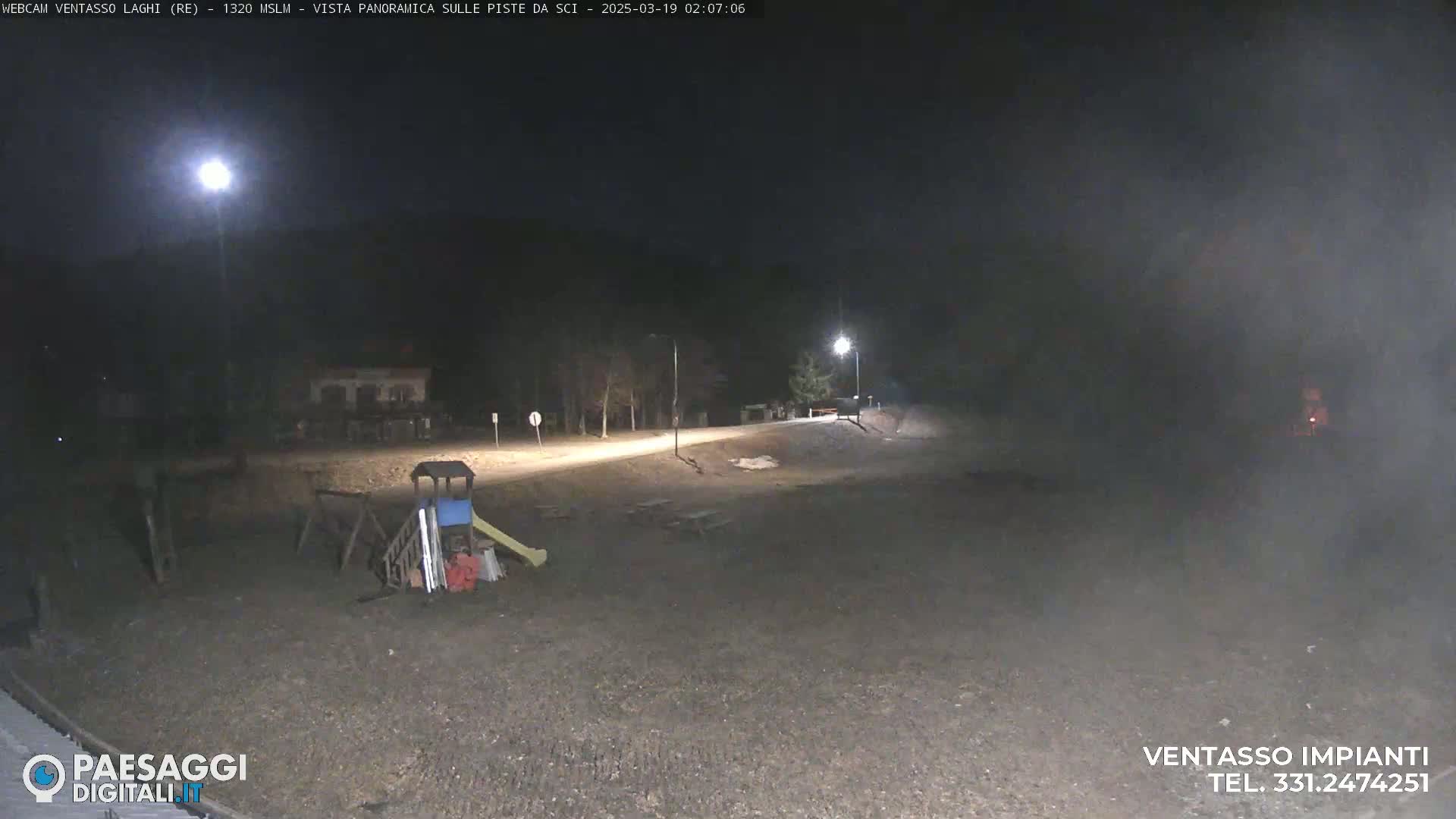 A nighttime, foggy view of an empty playground with a slide and climbing structure near a road, illuminated by streetlights.
