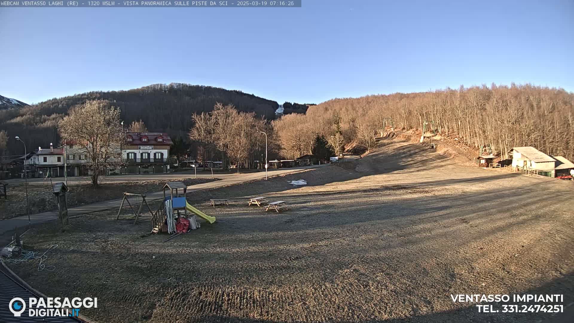 A mostly barren, brown ski slope is visible, with a playground, buildings, and leafless trees in the background under a clear blue sky.
