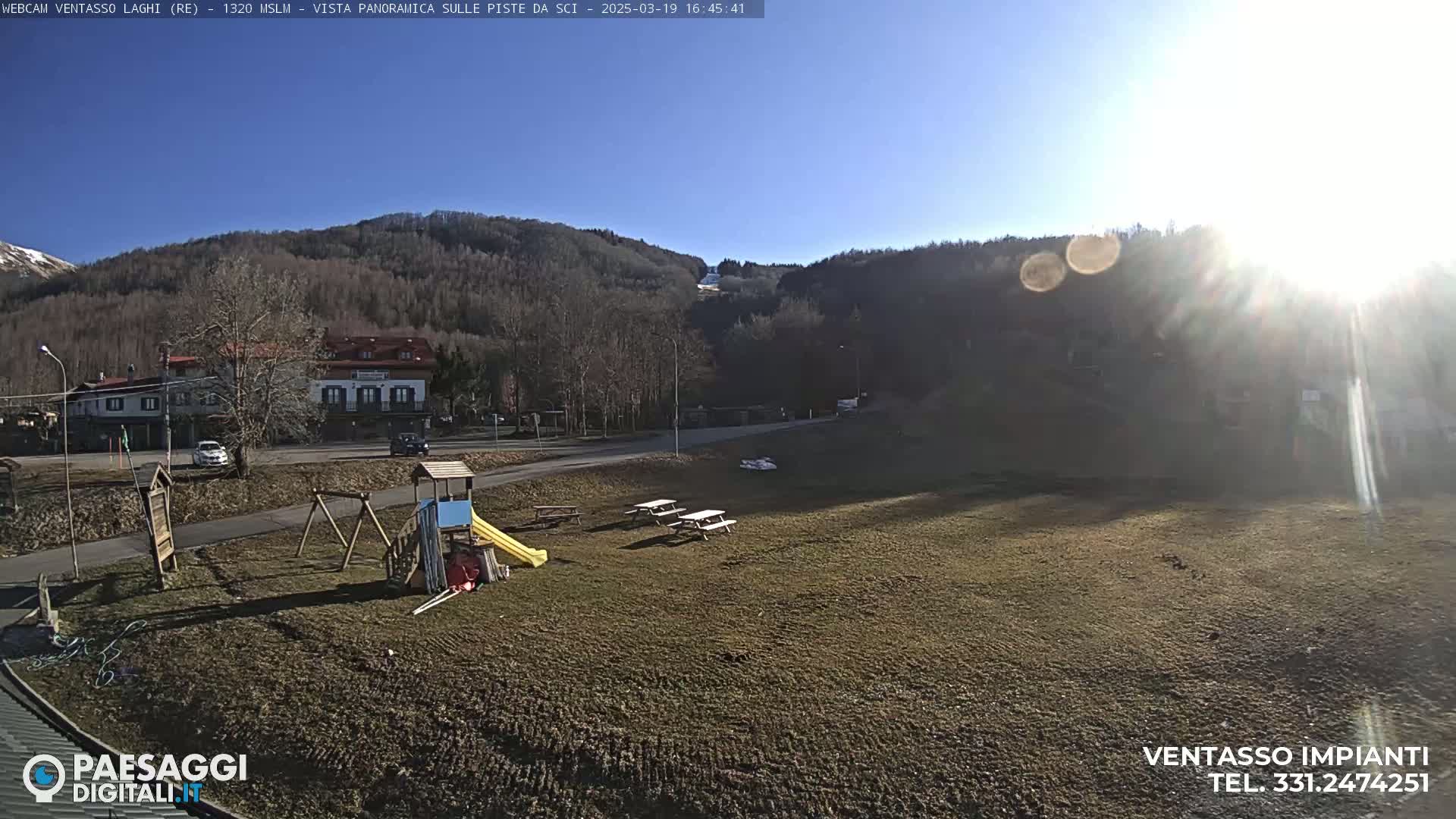 A sunny day reveals a playground, picnic tables, and buildings near a hillside covered in leafless trees.