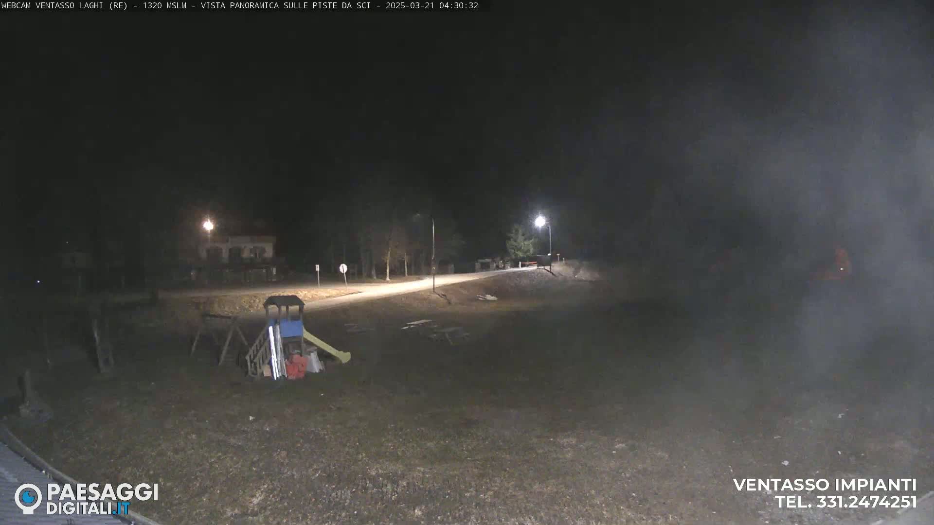 A nighttime, dimly lit view of an empty playground with a slide and climbing structures near a road, under a dark, possibly foggy sky.