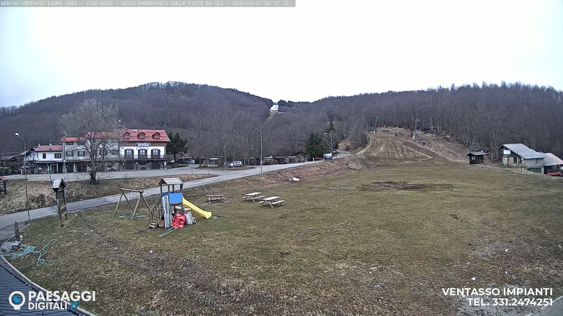 A mostly clear day reveals a ski slope in a mountainous region, with a small village, playground equipment, and picnic tables in the foreground.