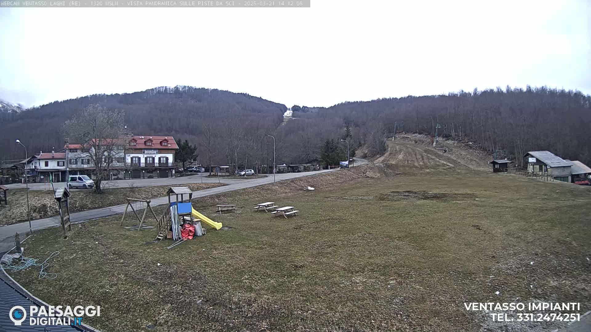 A mostly clear day reveals a village nestled at the base of a sparsely wooded hill with a ski slope visible in the background; a playground and picnic tables are in the foreground.