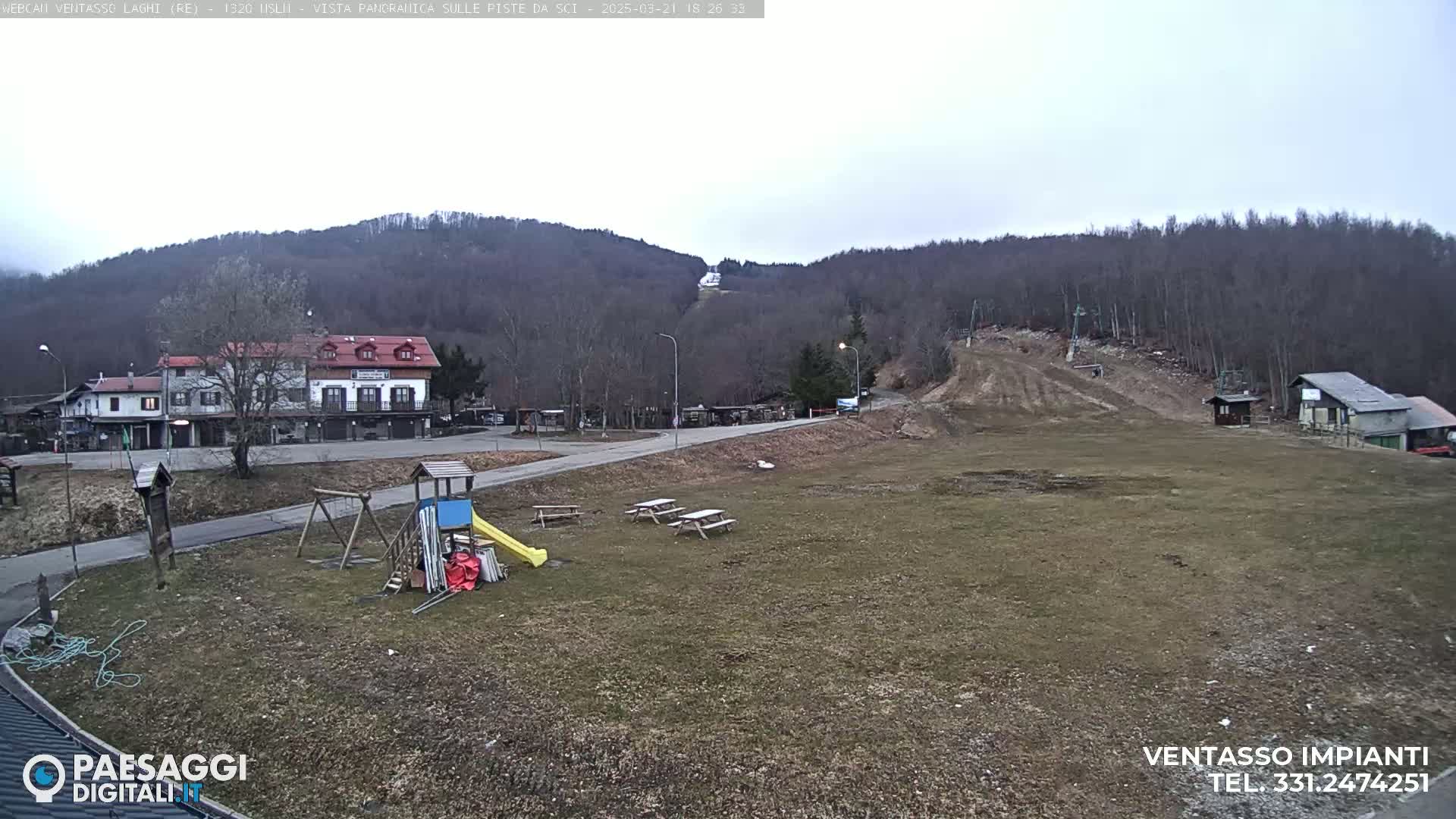 A view of a mostly barren, grassy area with a playground, picnic tables, and buildings in the background, under an overcast sky.