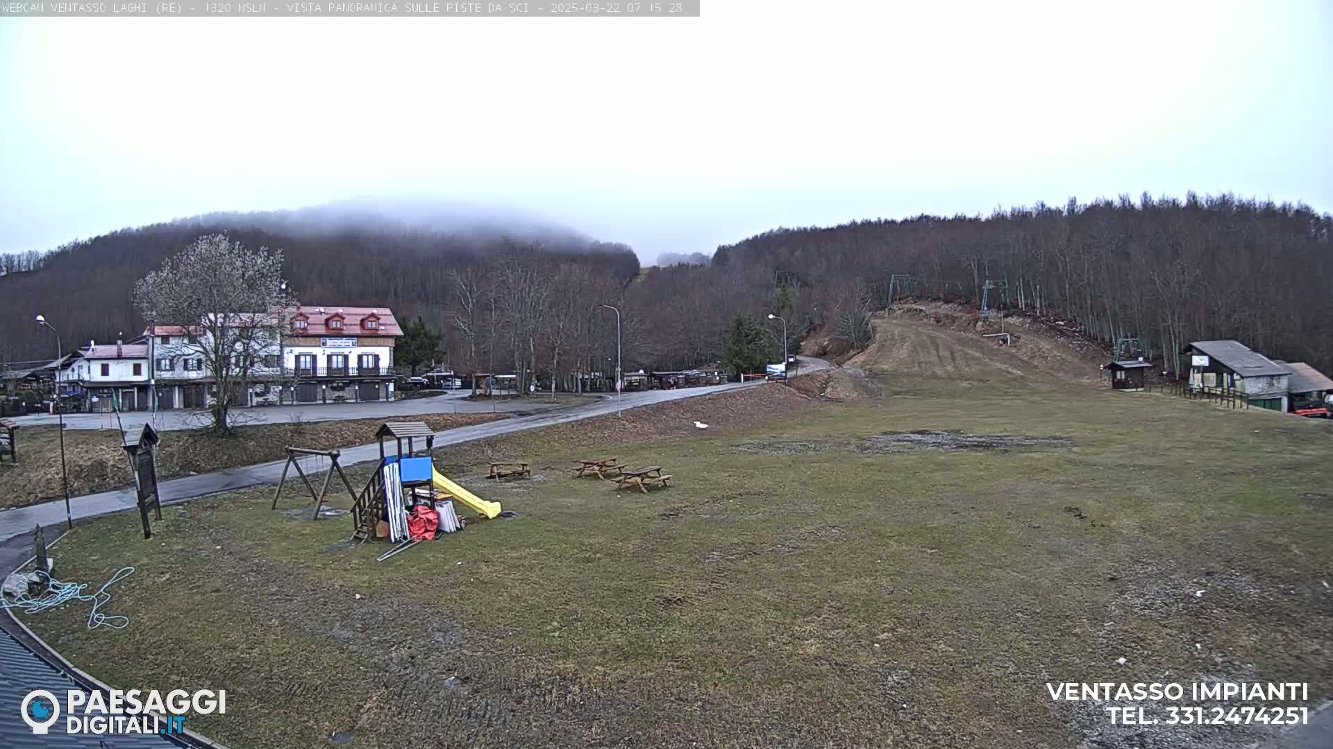 A playground and picnic tables sit in a grassy area below a ski slope, with buildings and a hill covered in fog in the background under an overcast sky.