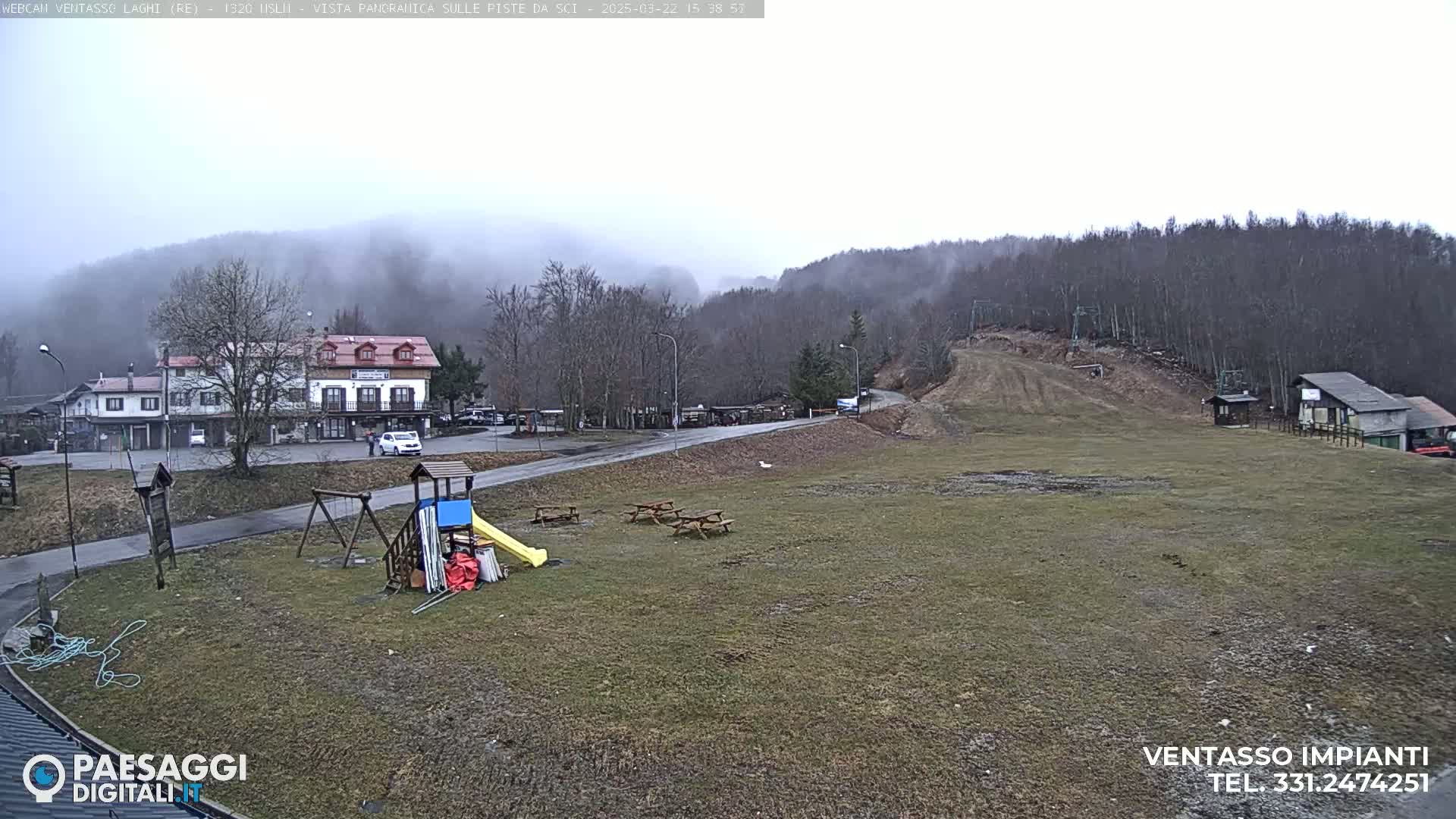 A mostly cloudy day reveals a grassy area with picnic tables, a playground, a building, and a ski lift in the background, all situated in front of a hill covered in leafless trees and partially obscured by fog.