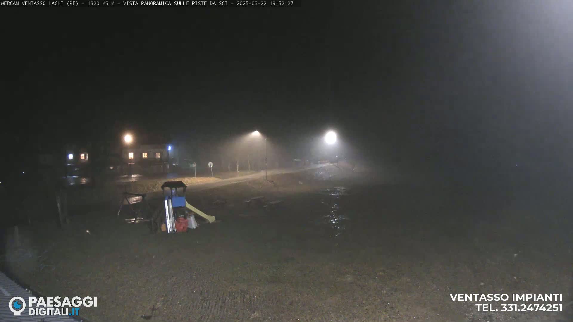 A nighttime, foggy view of a playground and a road near a building, illuminated by streetlights.