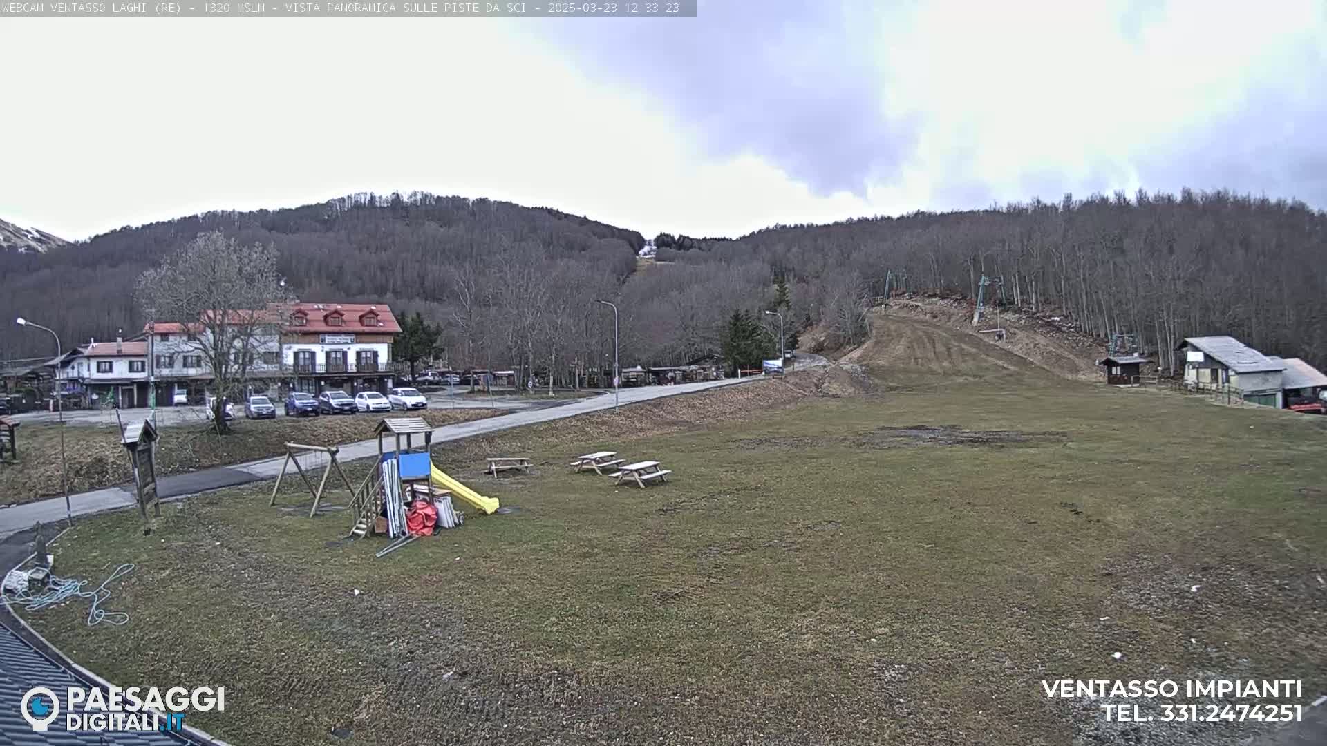 A mostly cloudy day reveals a view of a ski slope, a small playground, several picnic tables, a building, and parked cars, all set against a backdrop of bare trees on a hillside.