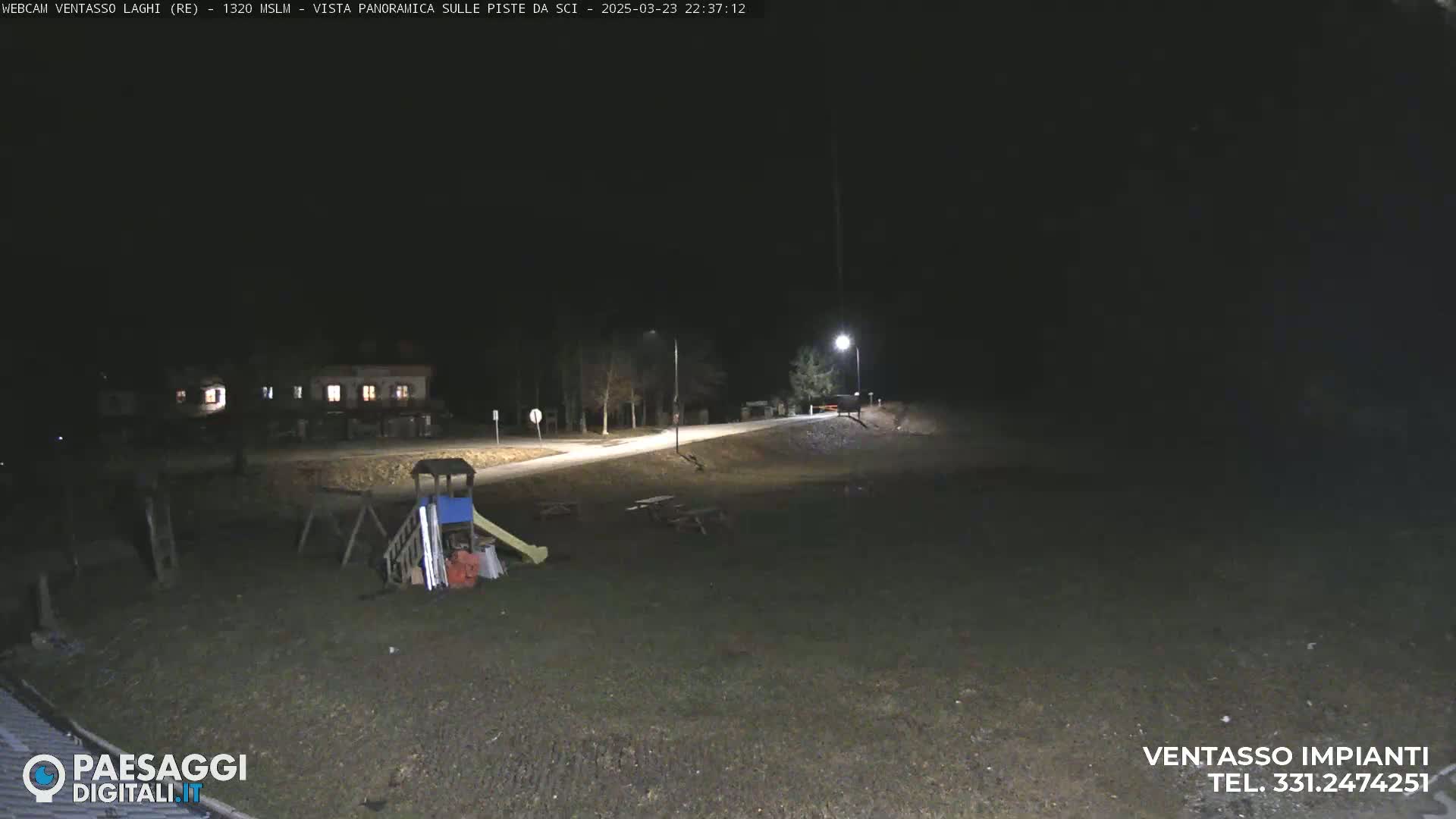 A nighttime view of an empty playground with a slide and climbing structure near a road, illuminated by a streetlight.