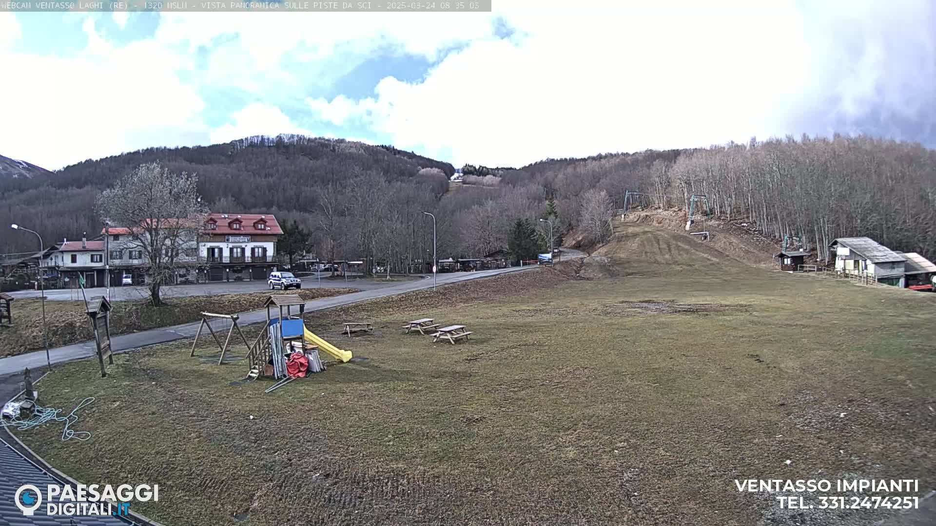 A mostly clear day reveals a grassy area with a playground, picnic tables, a small building, and a ski slope in the background, surrounded by leafless trees and hills.