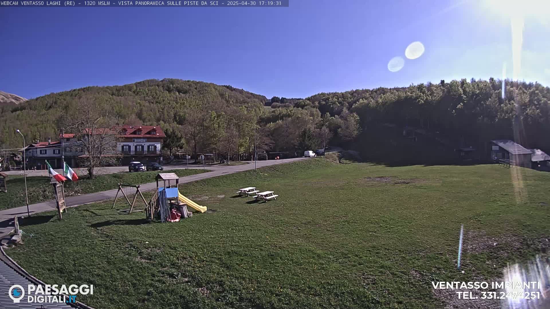 A playground and picnic tables sit on a grassy field in front of a building and wooded hillside under a sunny sky.