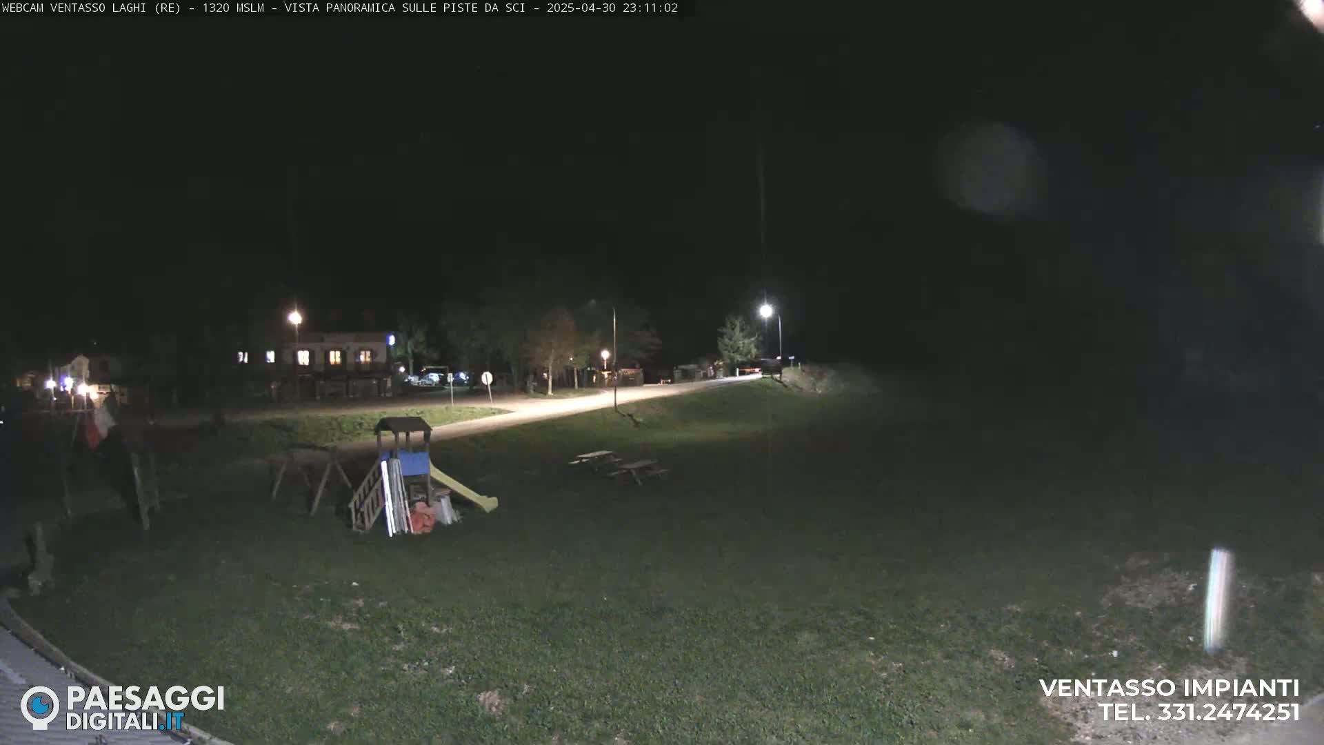 A nighttime view of a playground and picnic tables in a grassy area, with a road and buildings visible in the background under a dark sky.