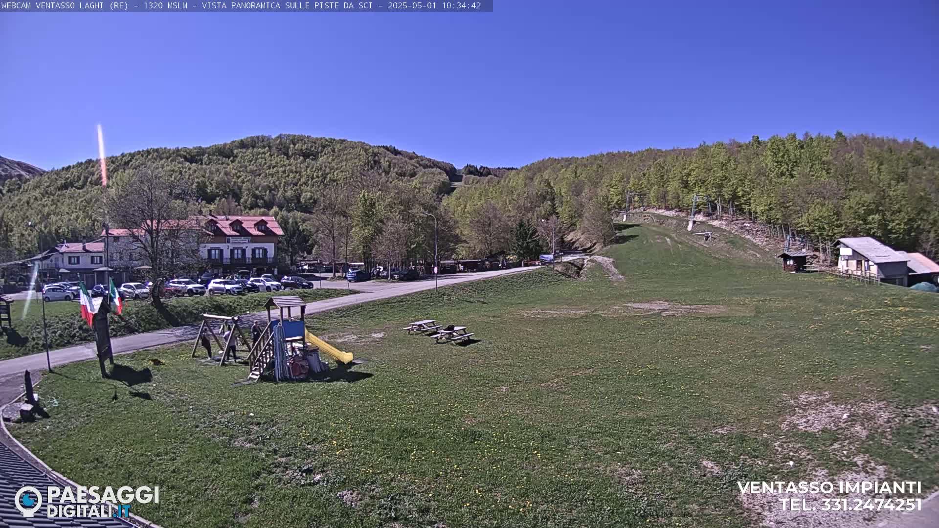 A sunny day in a mountainous area shows a parking lot, a playground, picnic tables, and ski lift infrastructure in a grassy field, with a building and forest in the background.