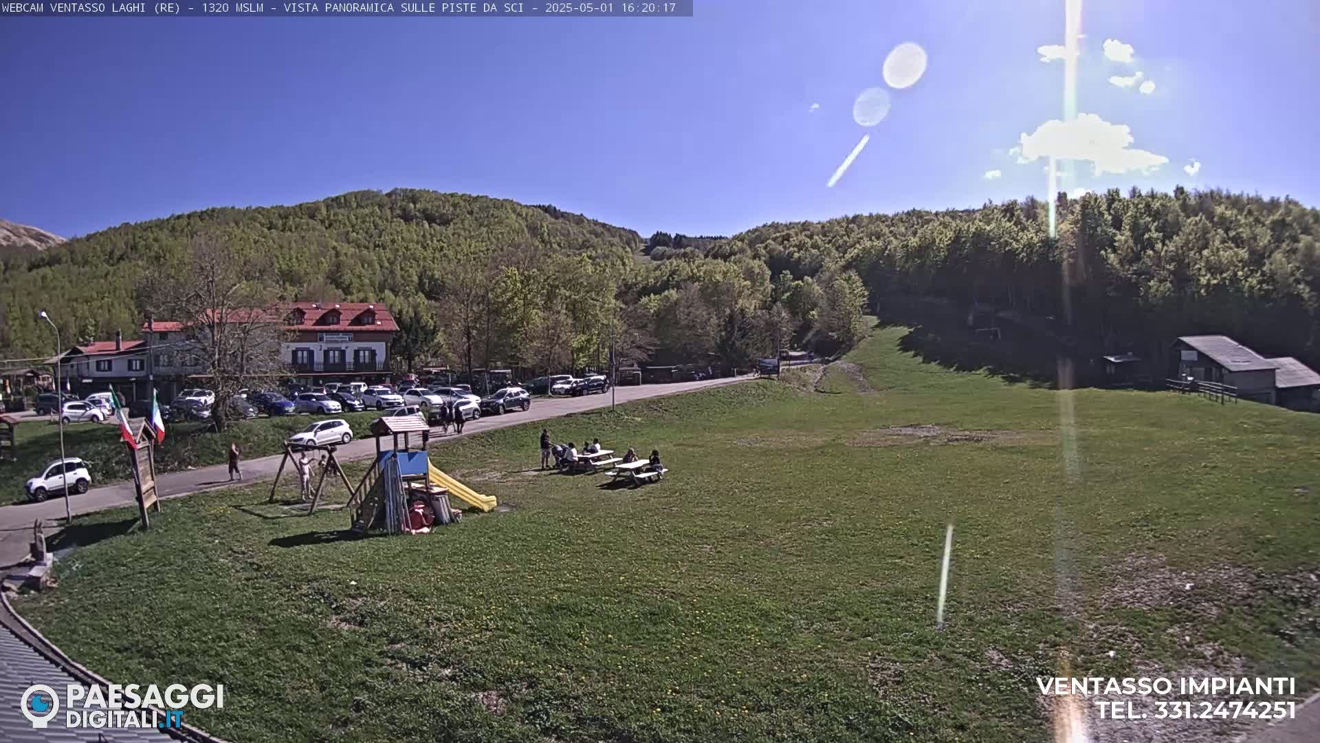 A sunny day at a mountain resort shows a parking lot full of cars near a building, a playground, and people sitting at picnic tables on a grassy field, all surrounded by green hills and trees.
